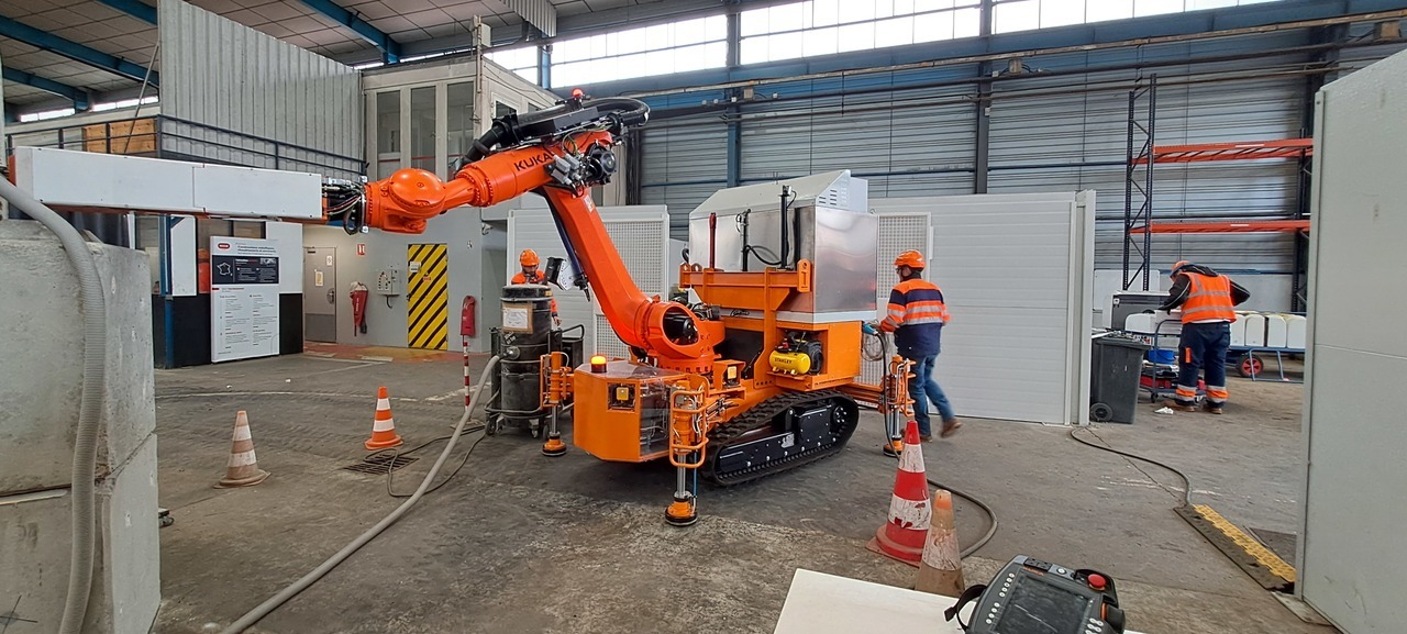 Un bras robotique KUKA orange sur une plateforme mobile à chenilles dans un atelier industriel, avec des ouvriers.