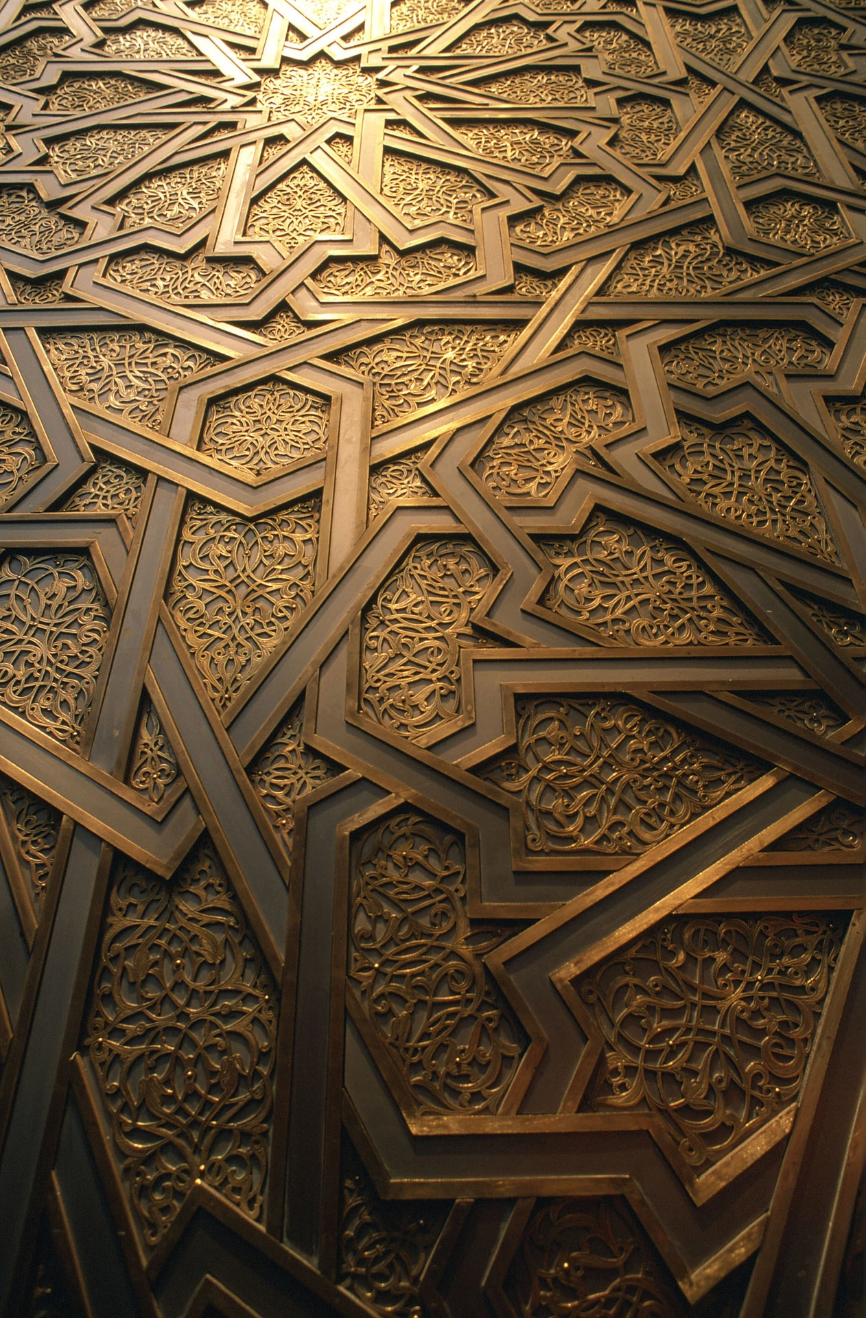Détail d'une surface décorative islamique en bronze et or, présentant des motifs géométriques entrelacés encadrant des arabesques florales d'un doré éclatant. La lumière incidente du haut accentue les reliefs et les reflets métalliques.