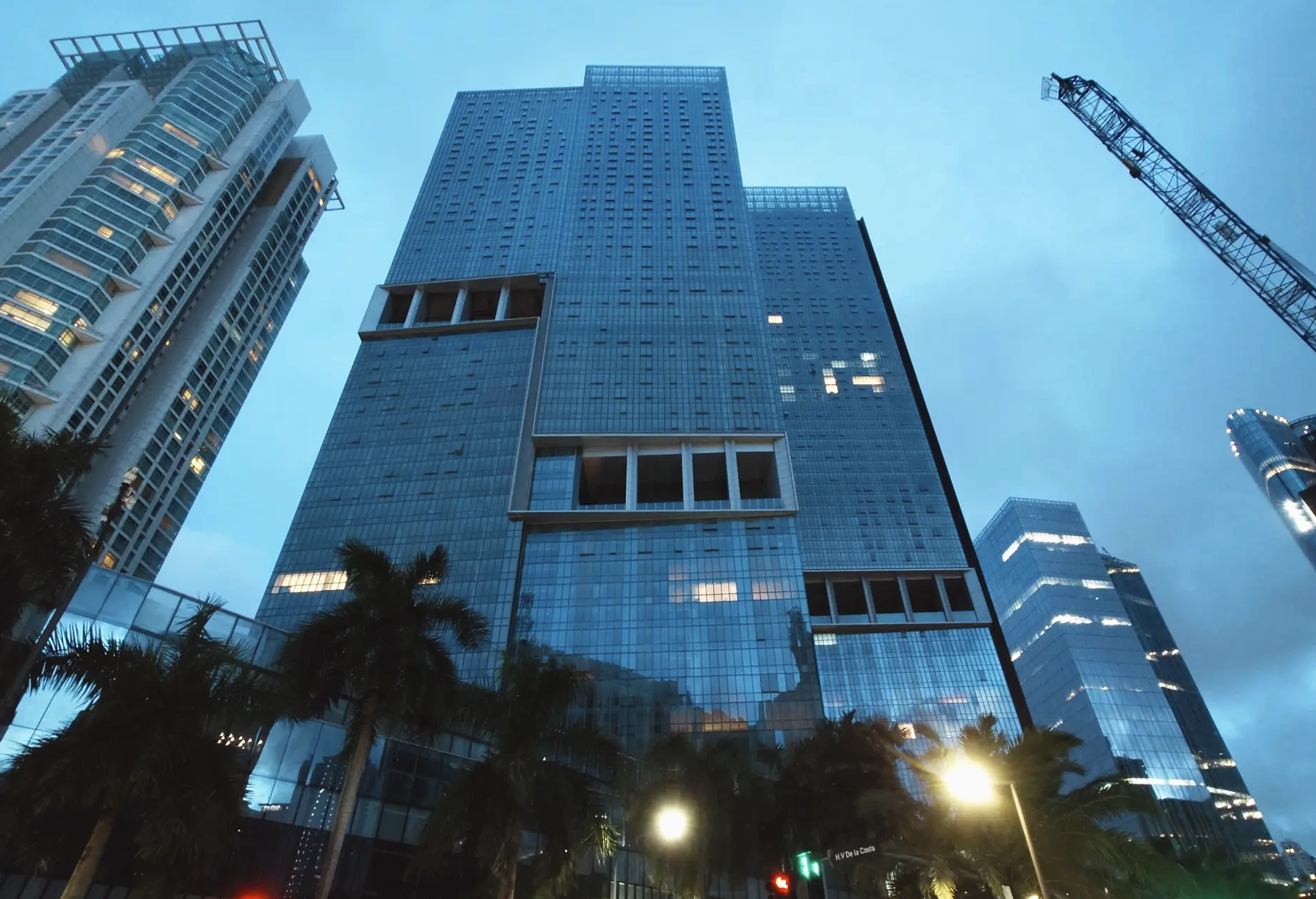 Plusieurs gratte-ciel modernes aux façades de verre bleutées se dressent contre un ciel de crépuscule sombre, leurs fenêtres éclairées reflétant des lumières urbaines et des palmiers au premier plan.