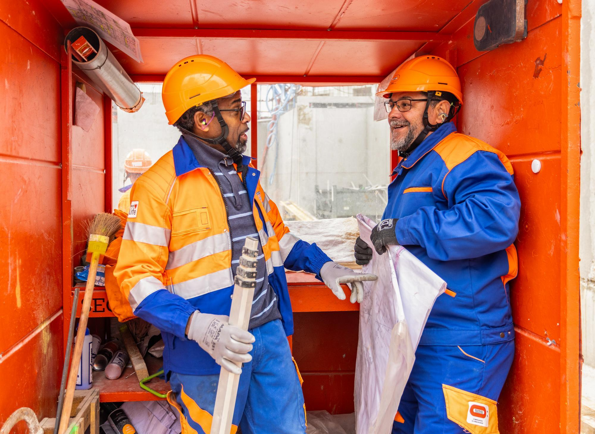 Deux ouvriers du bâtiment, portant des casques orange et des uniformes bleus et oranges, se parlent à l'intérieur d'une cabine de chantier orange vif, l'un tenant un plan et l'autre un jalon d'arpentage.