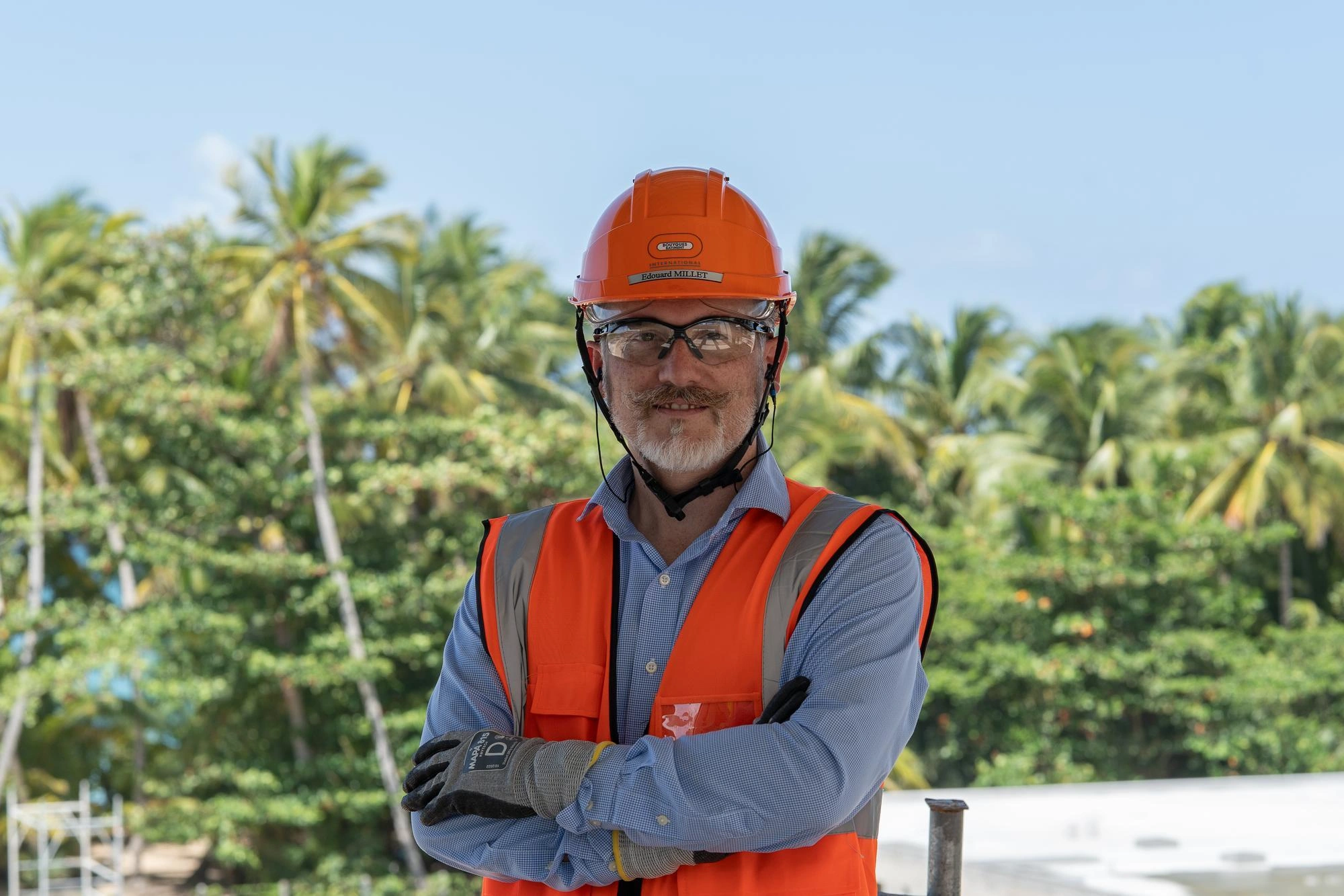 Portrait d'un homme souriant avec moustache et barbe, portant un casque de sécurité orange, des lunettes de protection, un gilet orange fluo et des gants de travail, les bras croisés, devant des palmiers et un ciel bleu.
