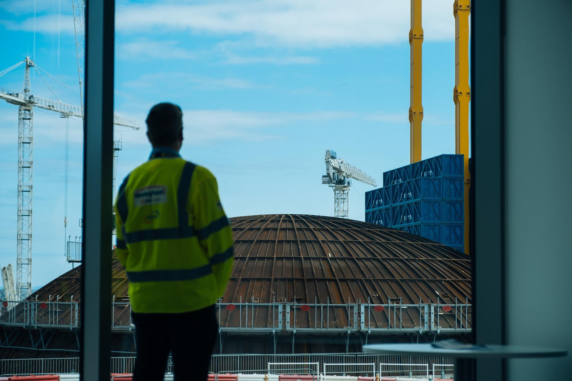 Une personne vêtue d'un gilet jaune haute visibilité est vue de dos, regardant à travers une fenêtre un chantier de construction sous un ciel bleu clair, avec de multiples grues blanches, une grande structure en dôme rouillée, des conteneurs bleus empilés et de hautes poutres jaunes.