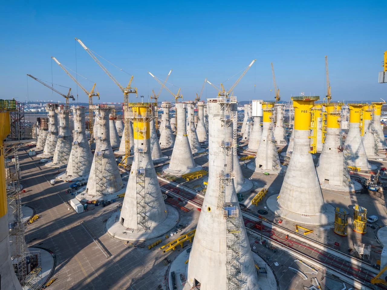 Vaste chantier montrant de nombreuses fondations coniques en béton gris en cours de construction, certaines avec des sommets jaunes vifs, et plusieurs grandes grues jaunes sous un ciel bleu dégagé.