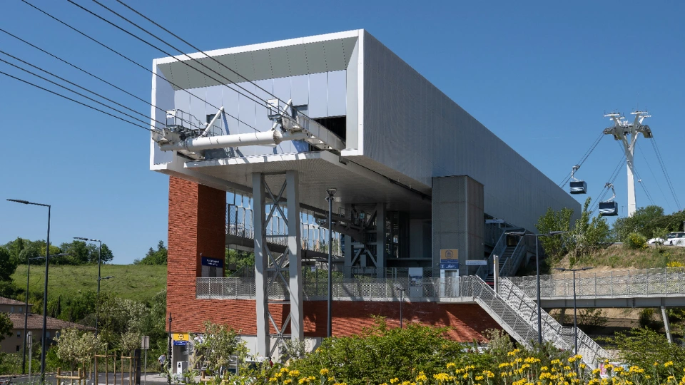 Gare de téléphérique moderne et élégante, avec un bâtiment en métal blanc et briques rouges, d'où partent des câbles supportant deux cabines sombres sous un ciel bleu. Le paysage environnant inclut des collines verdoyantes et des parterres de fleurs jaunes au premier plan.