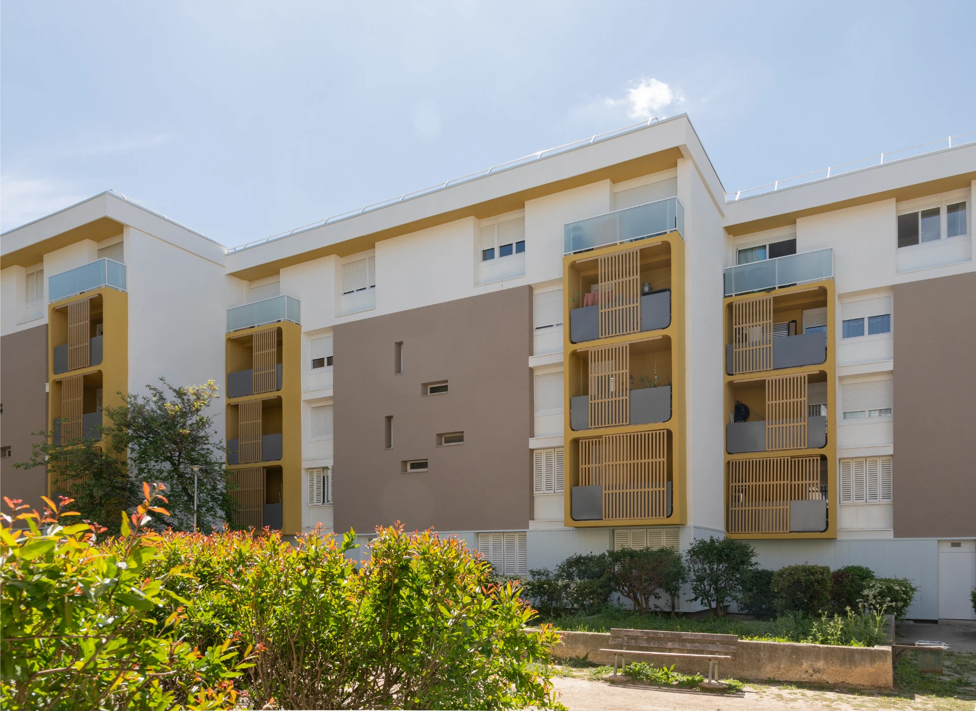 Modern apartment buildings with white and taupe facades, featuring numerous mustard yellow balconies with wooden slatted screens, set against a clear blue sky with green and red-leafed bushes in the foreground.