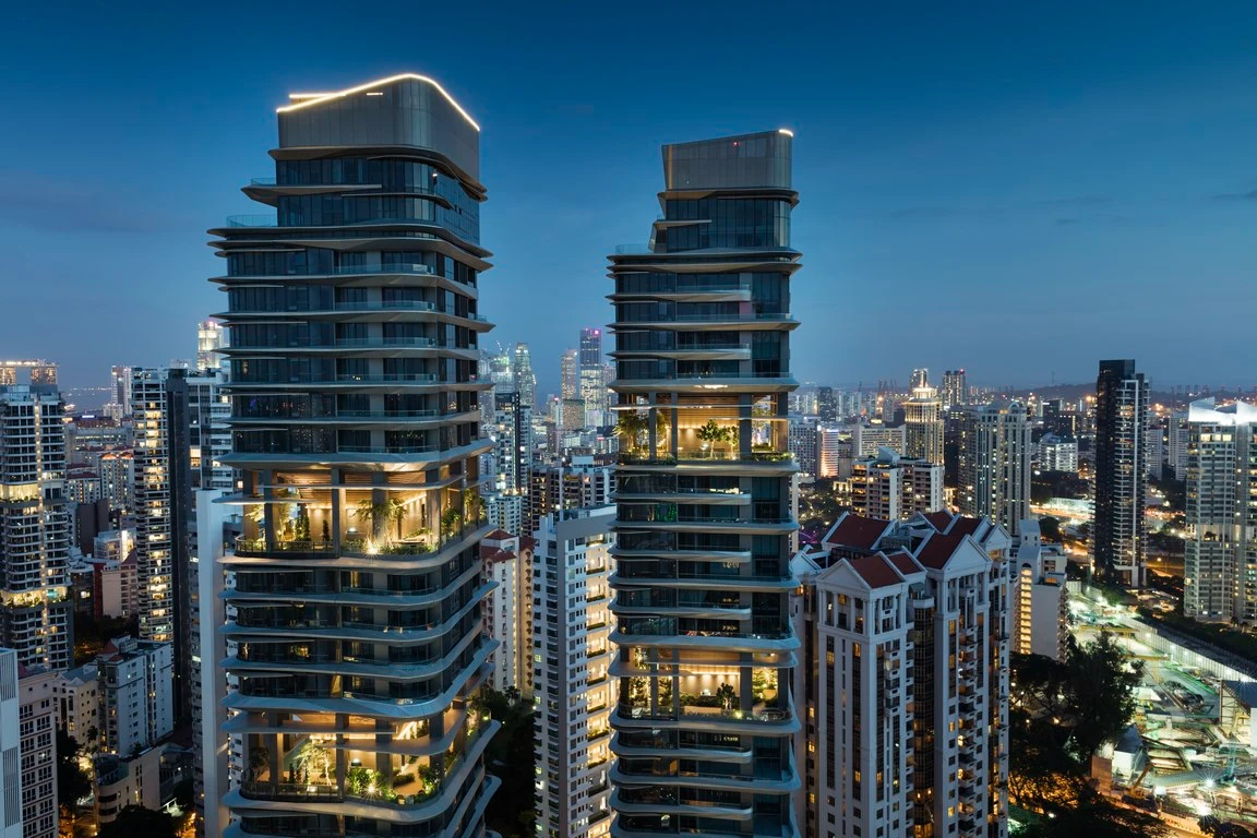 Deux gratte-ciel résidentiels modernes et stylisés, aux balcons incurvés et jardins illuminés, se dressent sous un ciel crépusculaire bleu foncé au milieu d'une ville tentaculaire illuminée.