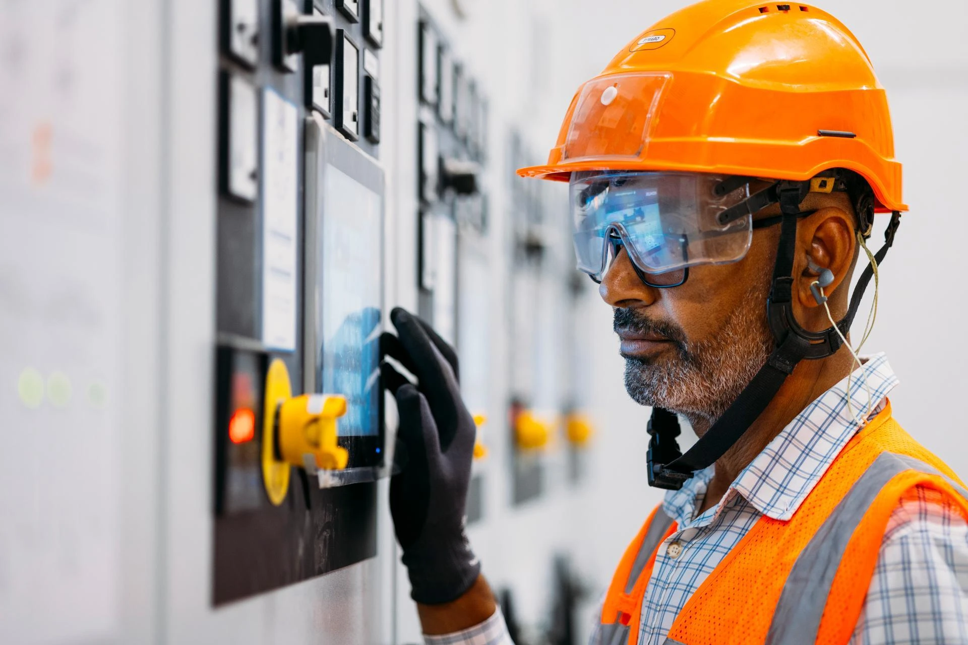 Un technicien aux cheveux gris, portant un casque de sécurité orange, des lunettes de protection et un gilet haute visibilité, manipule un écran tactile sur un tableau de commande industriel.