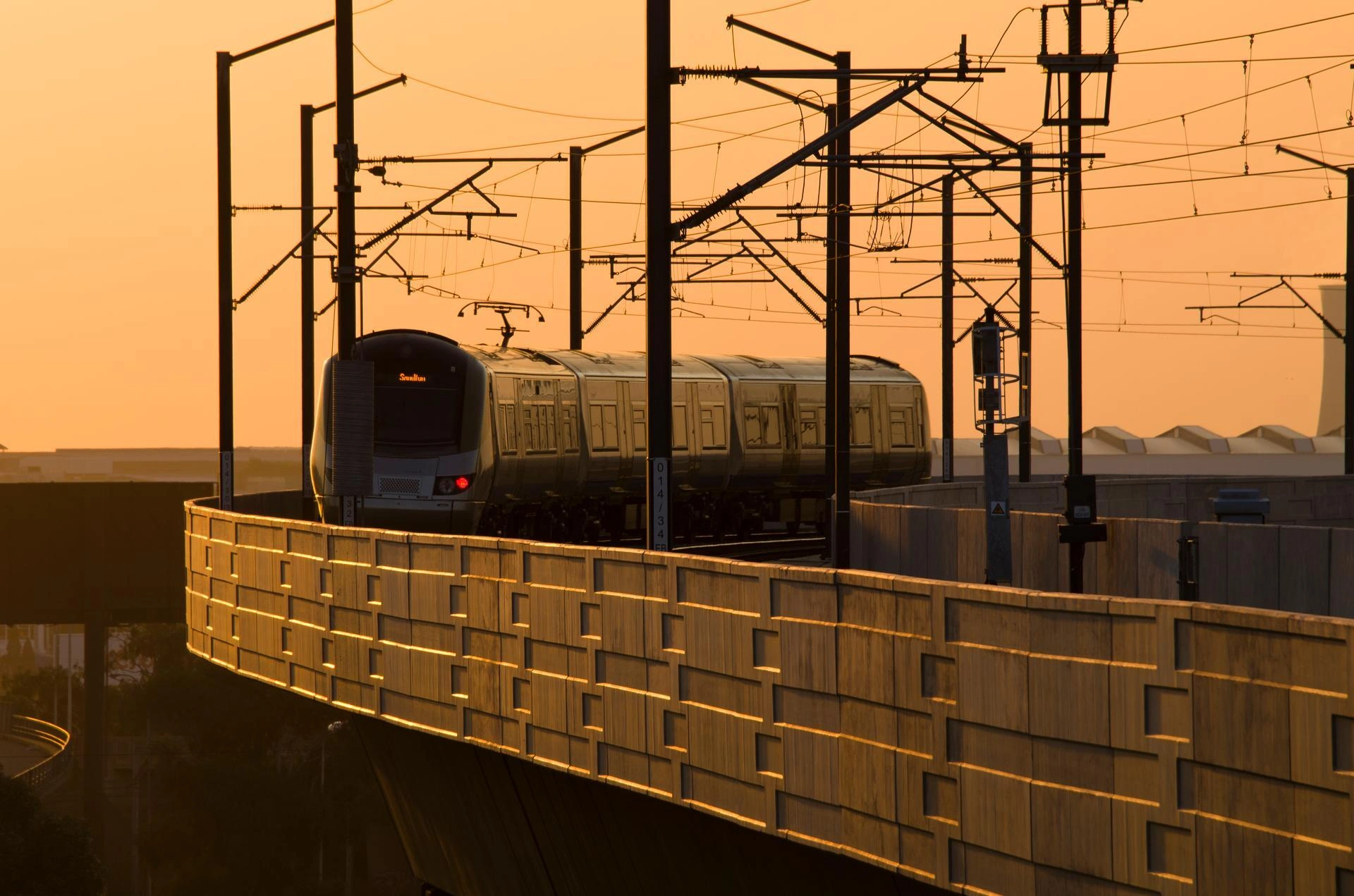 Un train moderne argenté et foncé circule sur un viaduc incurvé, baigné par la lumière dorée d'un coucher de soleil orange vif. Des pylônes et des caténaires se dessinent en silhouette nette sur le ciel lumineux.