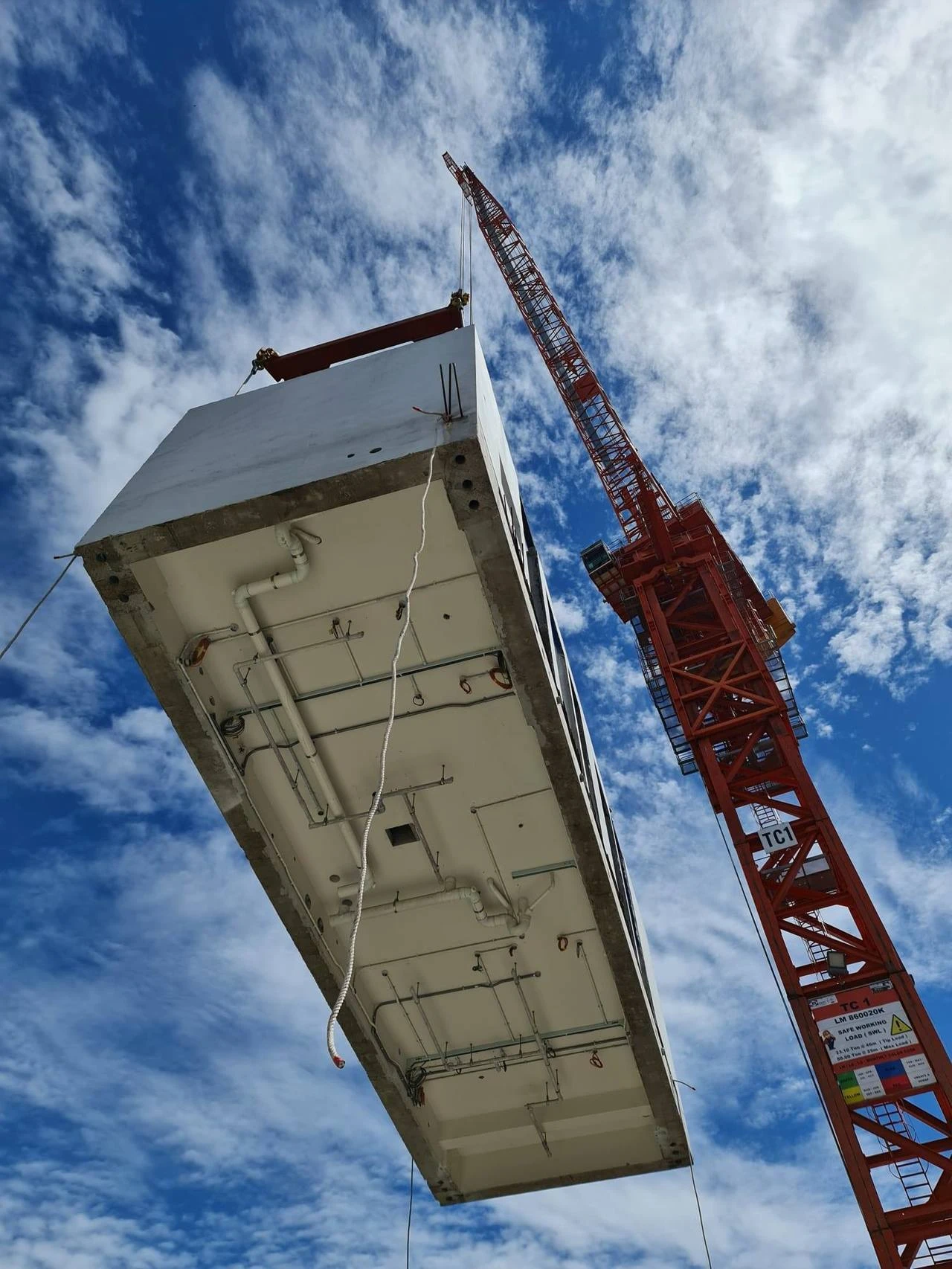 Grue rouge levant un module de construction préfabriqué en béton sous un ciel bleu nuageux.