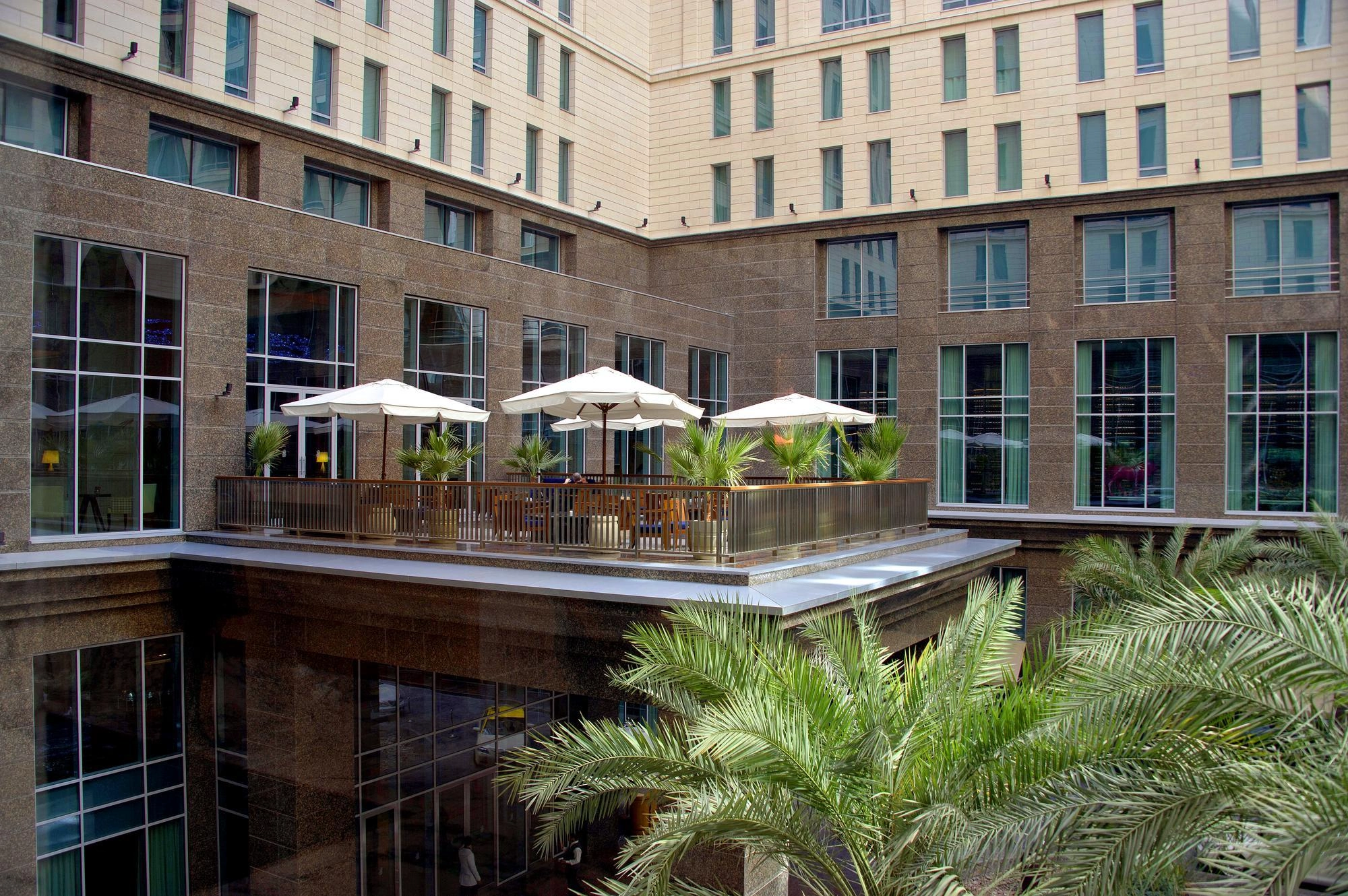 Terrasse extérieure avec parasols blancs et palmiers dans un bâtiment moderne à plusieurs étages.