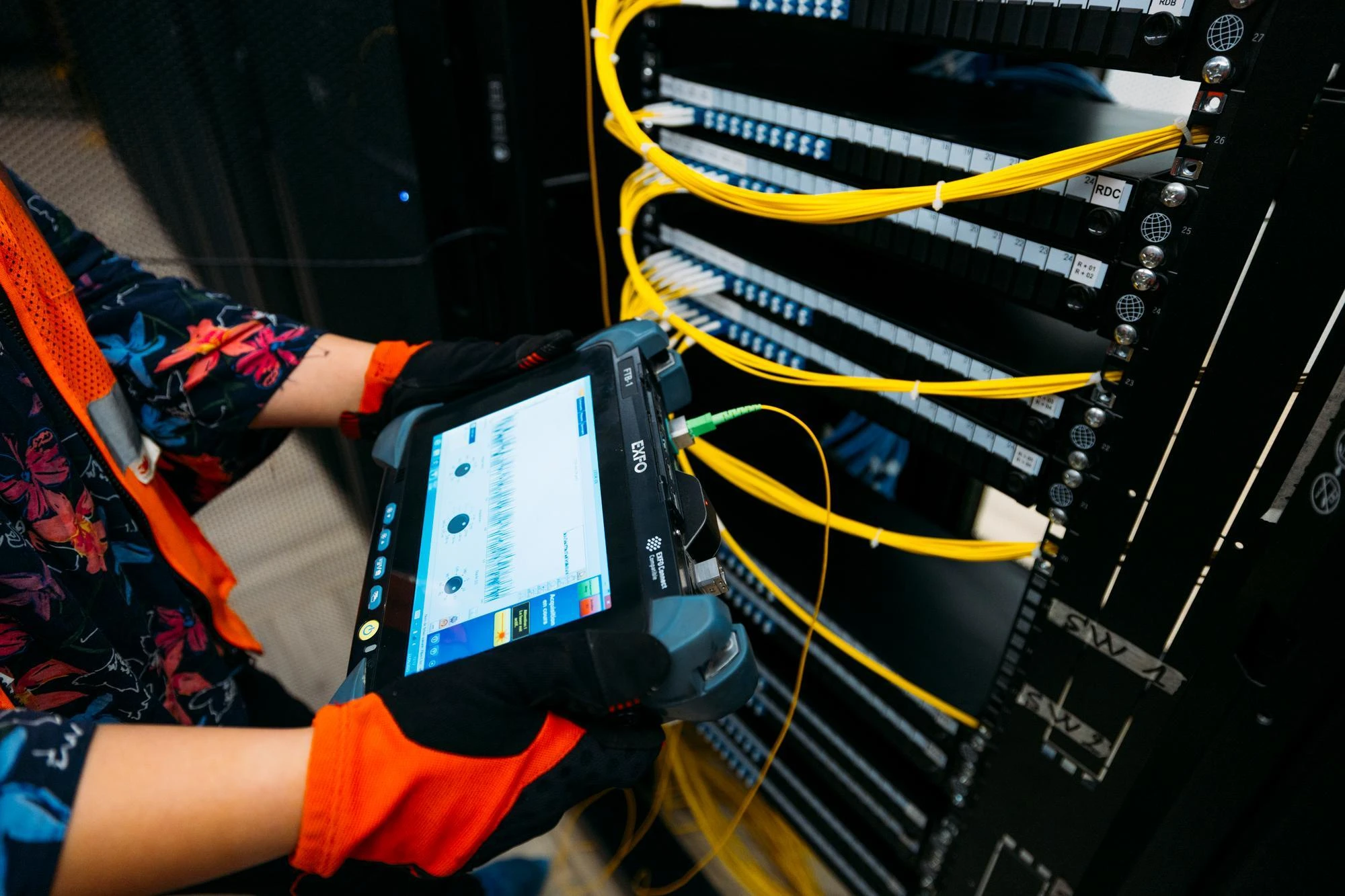 Mains gantées utilisant une tablette de test de fibre optique EXFO, connectée par un câble jaune à une baie de serveurs noire remplie de nombreux câbles de fibre optique jaunes vifs.