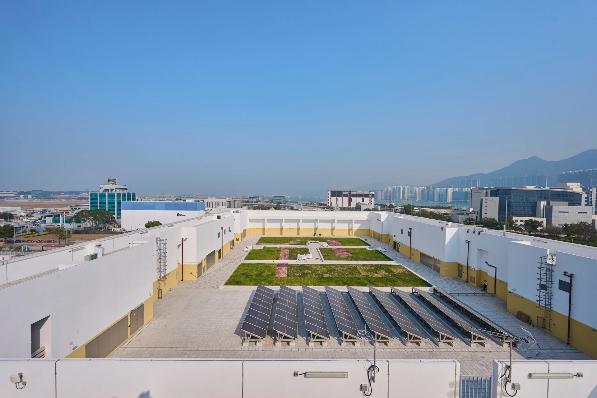 Vue aérienne d'un complexe de bâtiments modernes avec un toit-terrasse durable, comprenant un jardin verdoyant aux motifs floraux rouges et des rangées de panneaux solaires gris. L'arrière-plan révèle un paysage urbain étendu avec une tour de contrôle d'aéroport et des montagnes lointaines sous un ciel bleu clair mais brumeux.