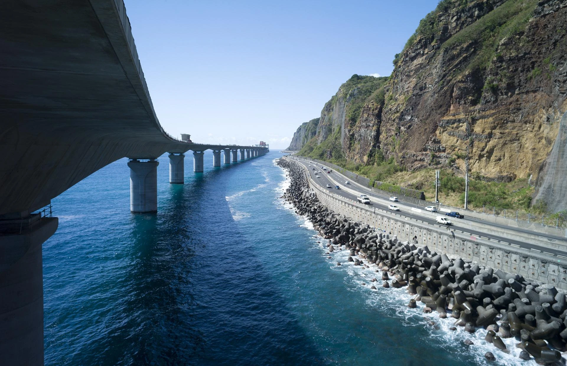 Un long viaduc en béton s'étend sur l'océan bleu profond à gauche, tandis qu'une autoroute côtière avec des voitures et des tétrapodes longe une falaise escarpée à droite, sous un ciel clair.