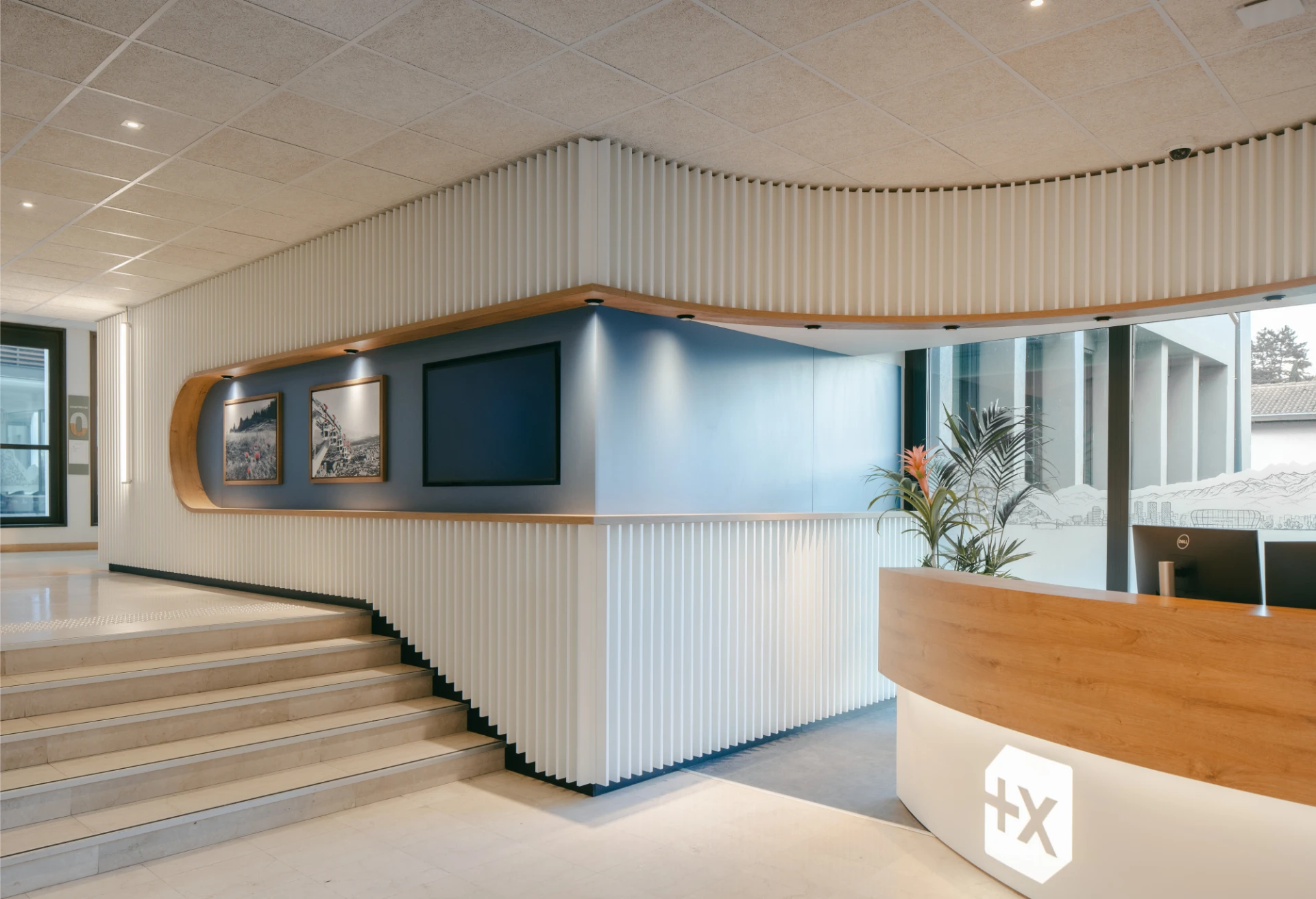 Réception moderne et élégante avec un comptoir d'accueil incurvé en bois clair et blanc, éclairé par un logo, devant un mur lamellé blanc qui intègre un panneau bleu avec des écrans et des cadres décoratifs. Des escaliers en pierre claire descendent à gauche, complétant l'ambiance lumineuse et professionnelle.