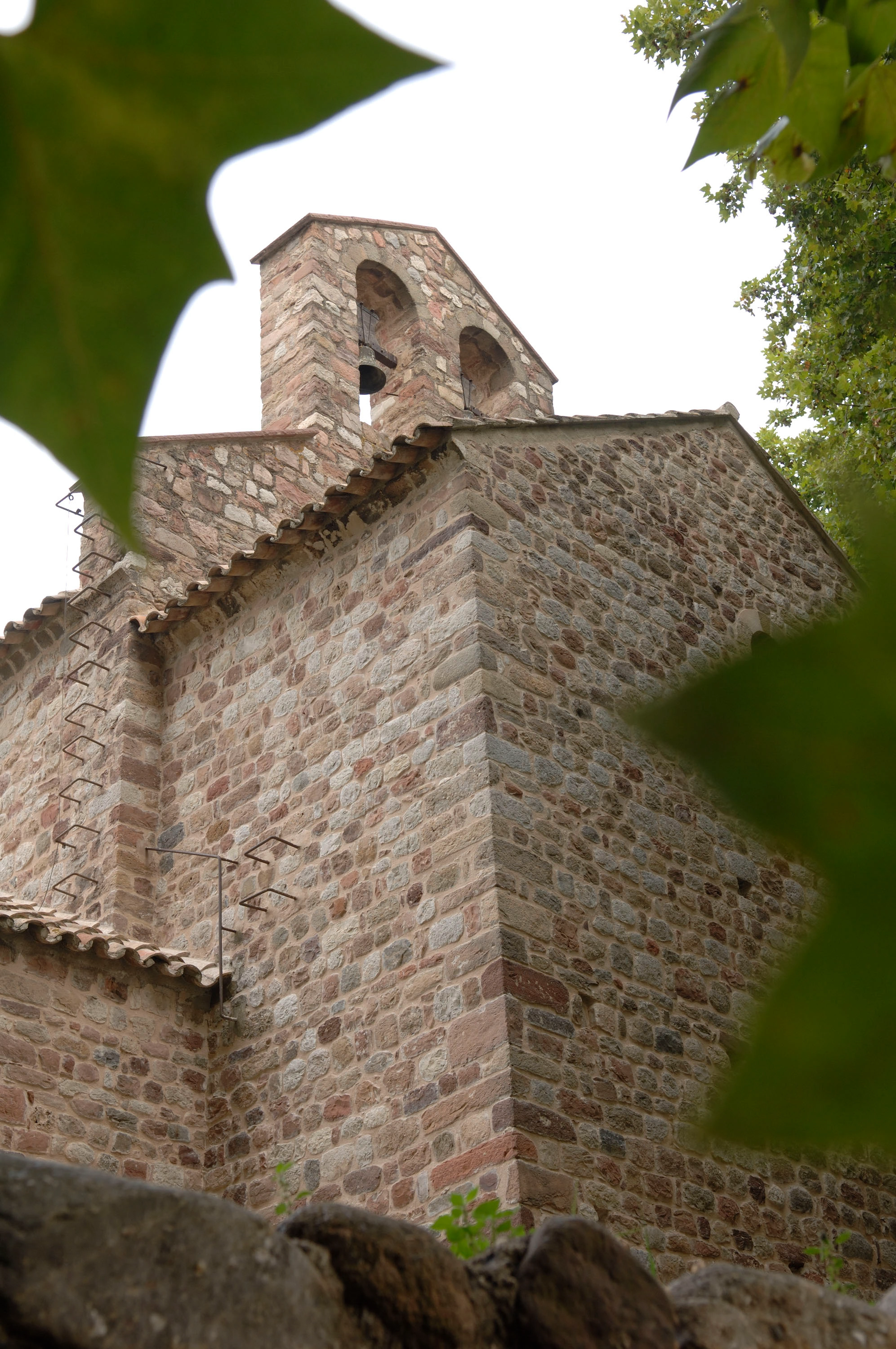 Une ancienne chapelle en pierre aux murs de moellons bruns et gris, dotée d'un clocher à deux arcades avec une cloche et d'un toit de tuiles en terre cuite, est partiellement encadrée par des feuilles vertes en premier plan sur un ciel clair.