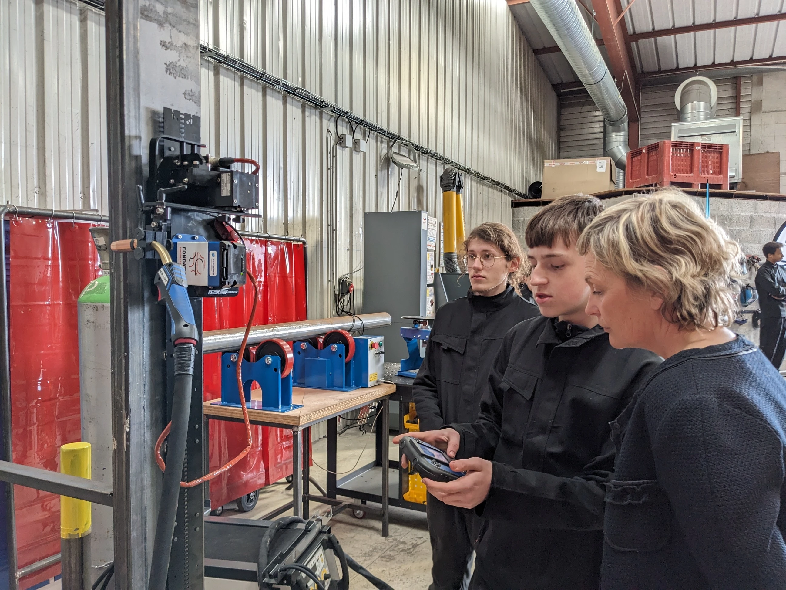 Trois personnes, deux jeunes hommes en uniforme noir et une femme aux cheveux blonds, examinent un poste de soudage industriel avec une torche et une pièce métallique sur des supports bleus. Un des jeunes hommes tient une télécommande, et des rideaux de protection rouges sont visibles en arrière-plan dans l'atelier.