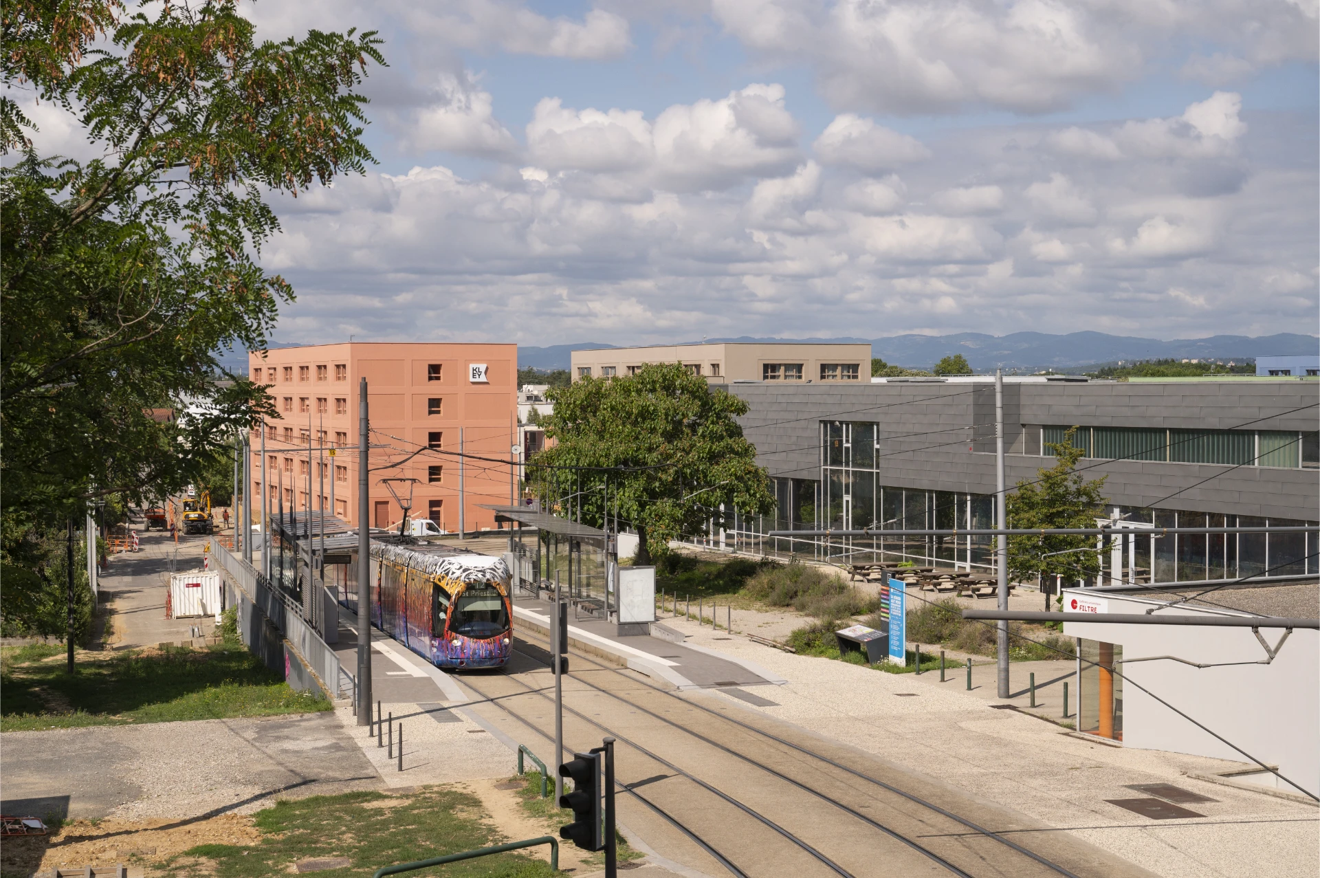 Un tramway multicolore et graffité est arrêté à une station moderne, encadré par un bâtiment couleur pêche à gauche et un bâtiment gris à droite sous un ciel parsemé de nuages blancs. Le premier plan est bordé de verdure et on aperçoit des travaux sur la gauche, avec des montagnes à l'horizon.