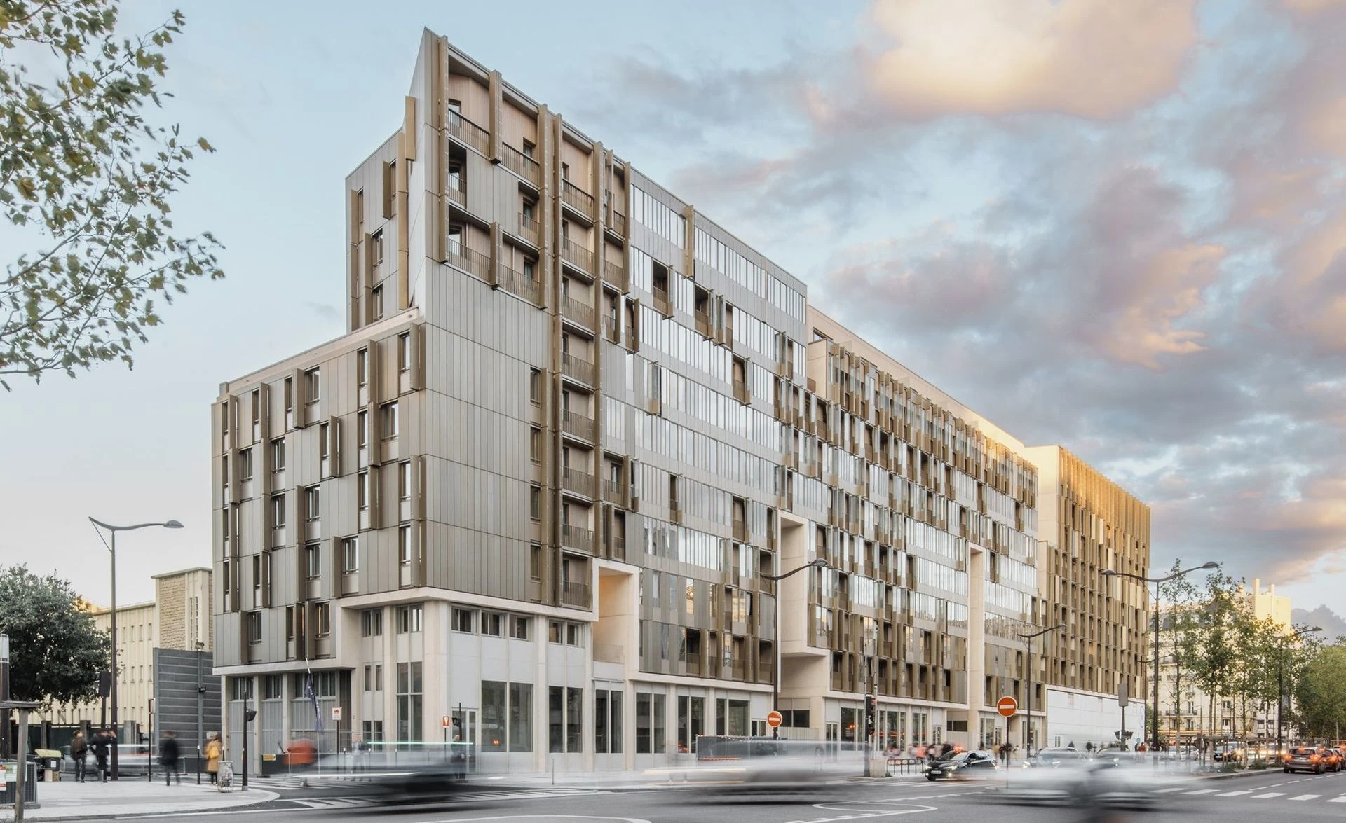 Un grand bâtiment moderne aux façades complexes mêlant panneaux bronze et fenêtres réfléchissantes s'élève le long d'une intersection urbaine animée, sous un ciel nuageux teinté de rose où les lumières des voitures en mouvement sont floues.