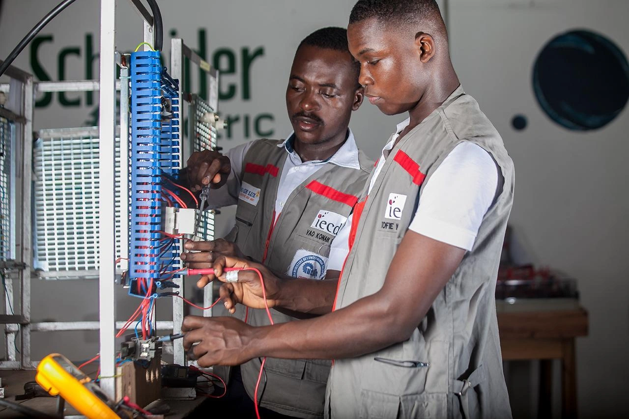 Deux hommes à la peau foncée en gilets de travail gris et rouges examinent un panneau électrique avec des borniers bleus et des fils colorés, l'un utilisant un tournevis et l'autre tenant des sondes de multimètre, dans un contexte de formation technique.