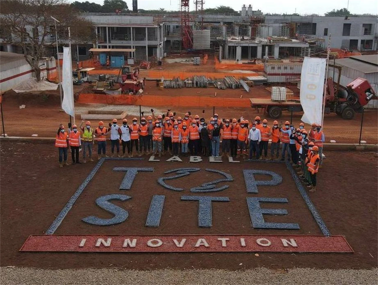 Un grand groupe de travailleurs en gilets et casques de sécurité orange posent sur un vaste chantier de construction. Ils se tiennent derrière une grande inscription au sol "LABEL TOP SITE INNOVATION" en lettres grises et rouges, avec des bâtiments en construction en arrière-plan.