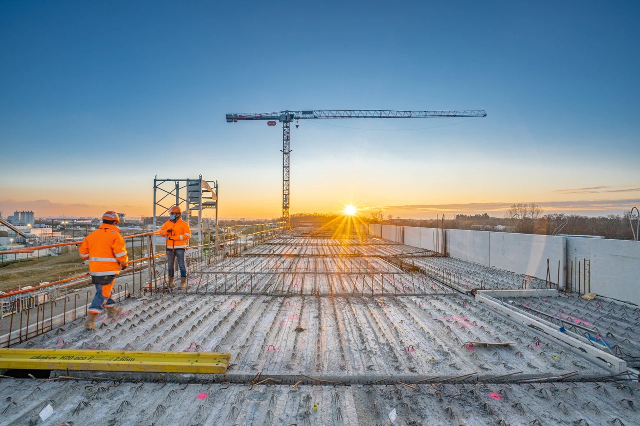 Deux ouvriers du bâtiment en tenue orange se tiennent sur une grande dalle de béton armé en construction, avec un coucher de soleil éclatant à l'horizon et une grue de chantier en arrière-plan.