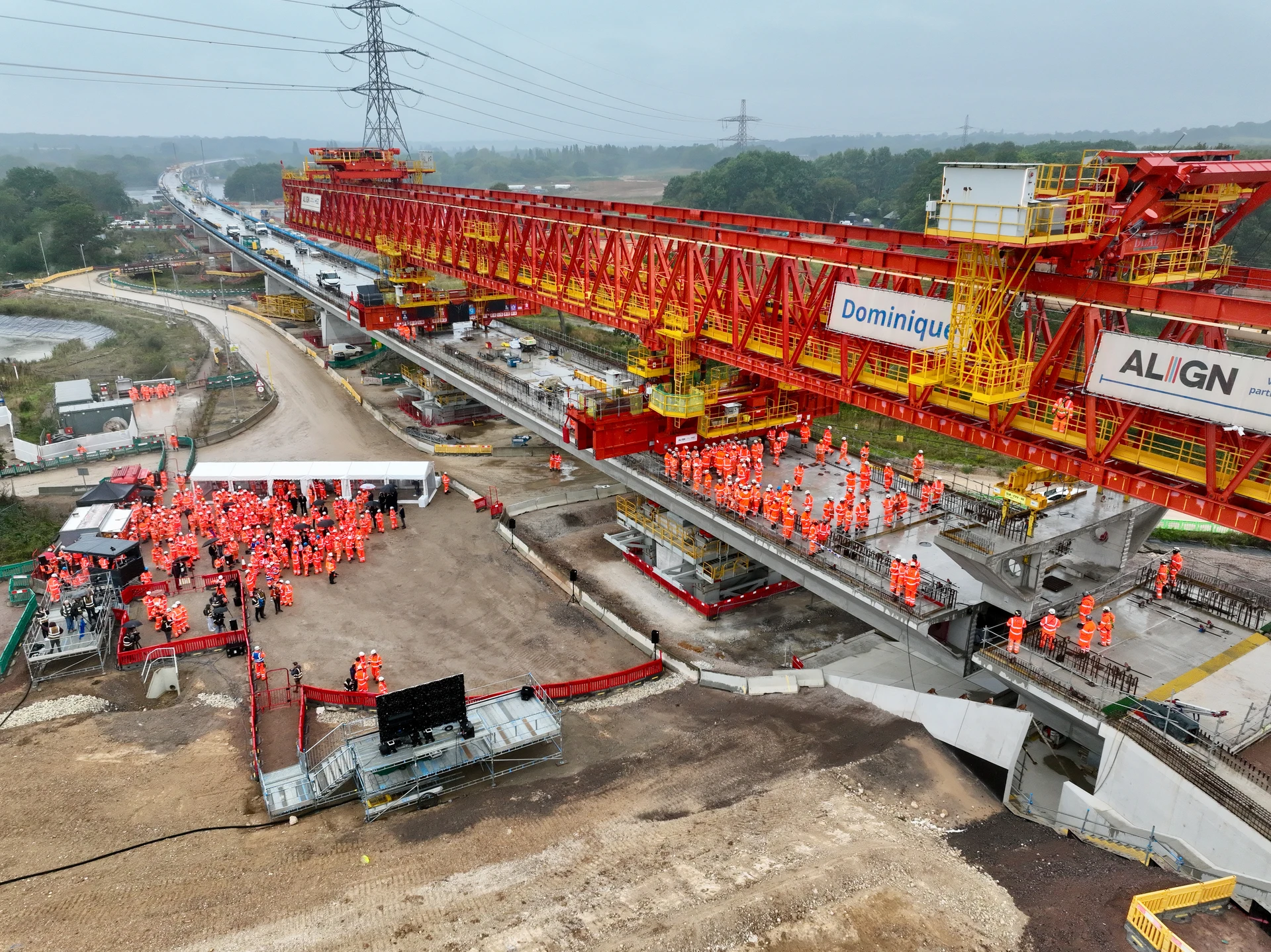 Vue aérienne d'un immense portique de lancement rouge et jaune, nommé "Dominique", construisant un viaduc sur un vaste chantier. De nombreux ouvriers en tenues orange haute-visibilité sont rassemblés sur la structure et au sol, sous un ciel gris.