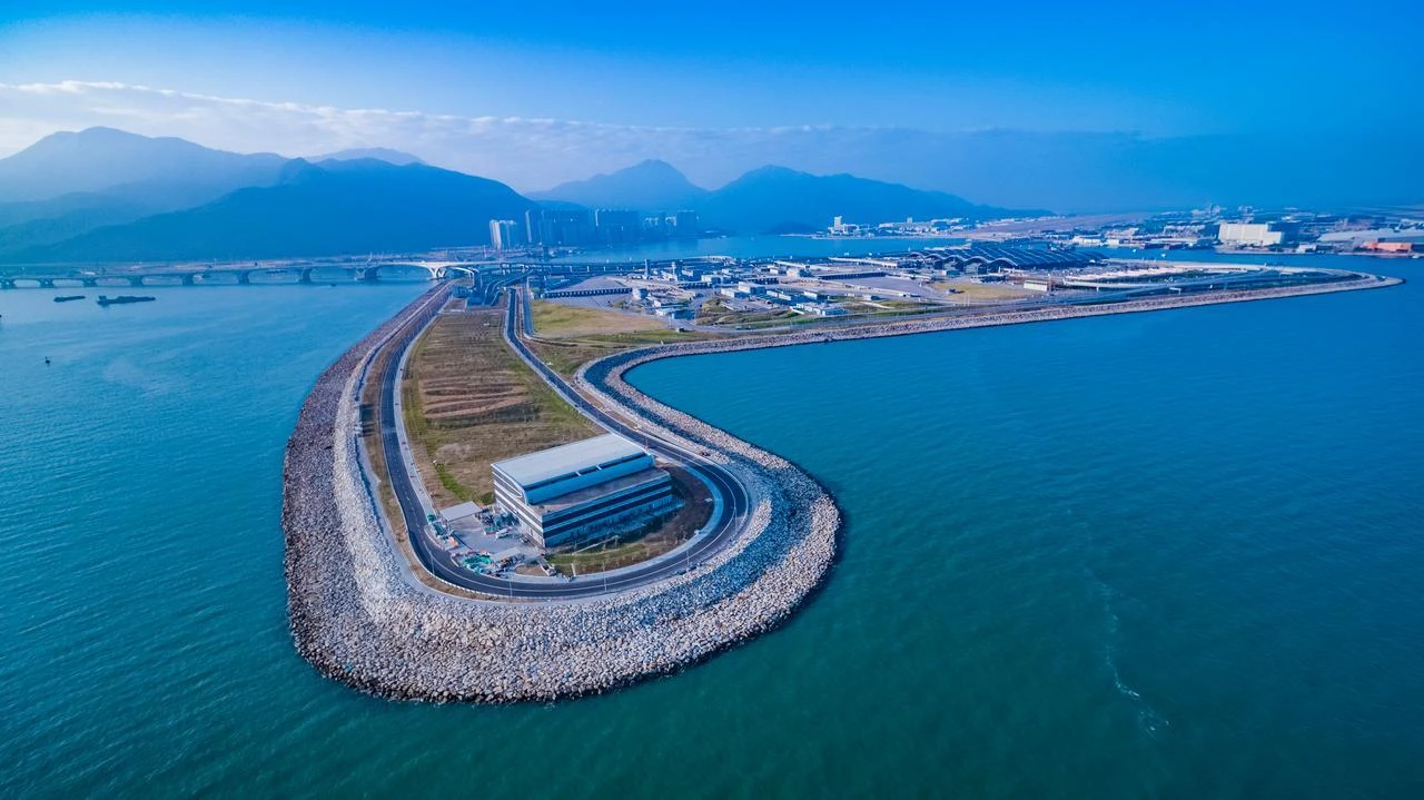 Vue aérienne d'une île artificielle courbée avec un bâtiment moderne et des routes, s'étendant dans la mer d'un bleu profond vers un aéroport et des montagnes lointaines sous un ciel clair.