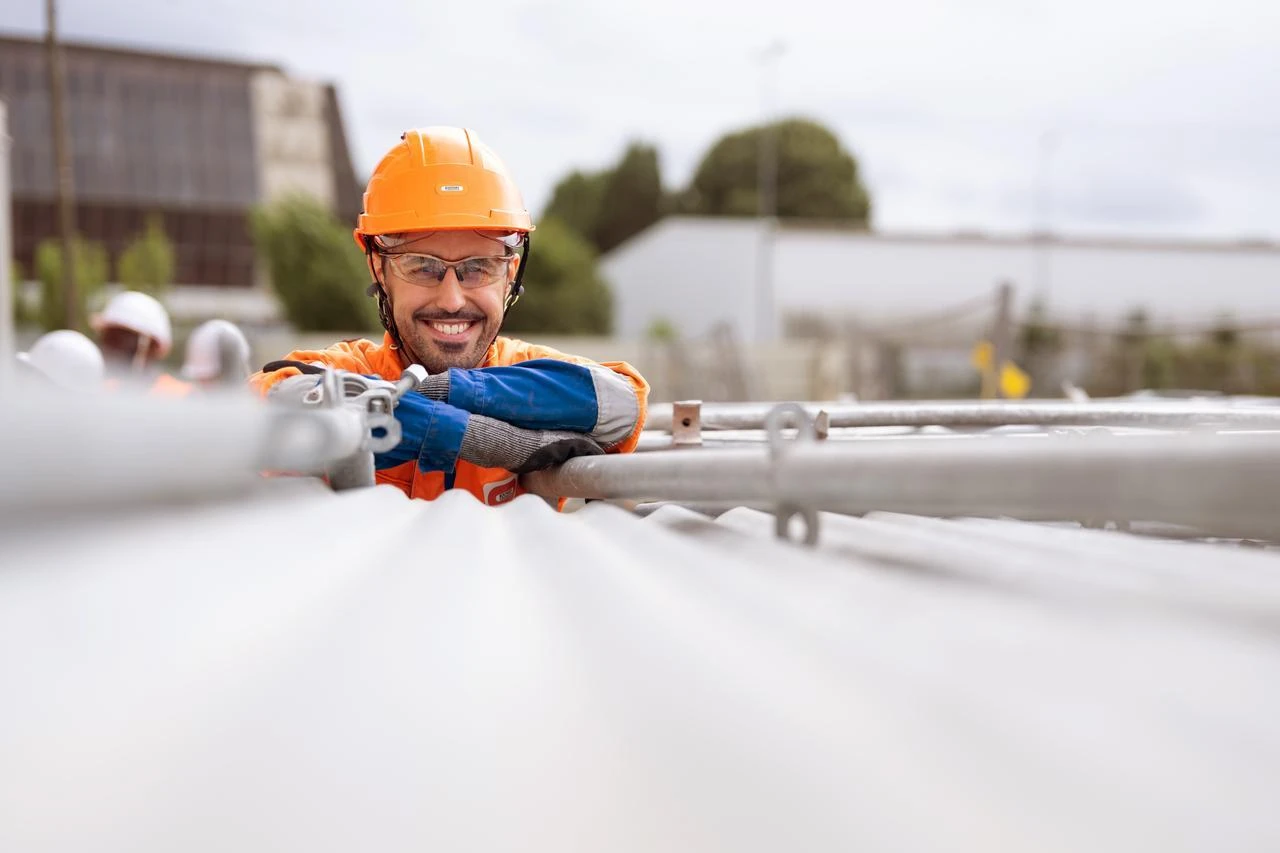 Un travailleur souriant en casque de sécurité orange et lunettes de protection, vêtu d'une tenue de travail orange et bleue, se penche sur des structures métalliques sur un chantier extérieur.