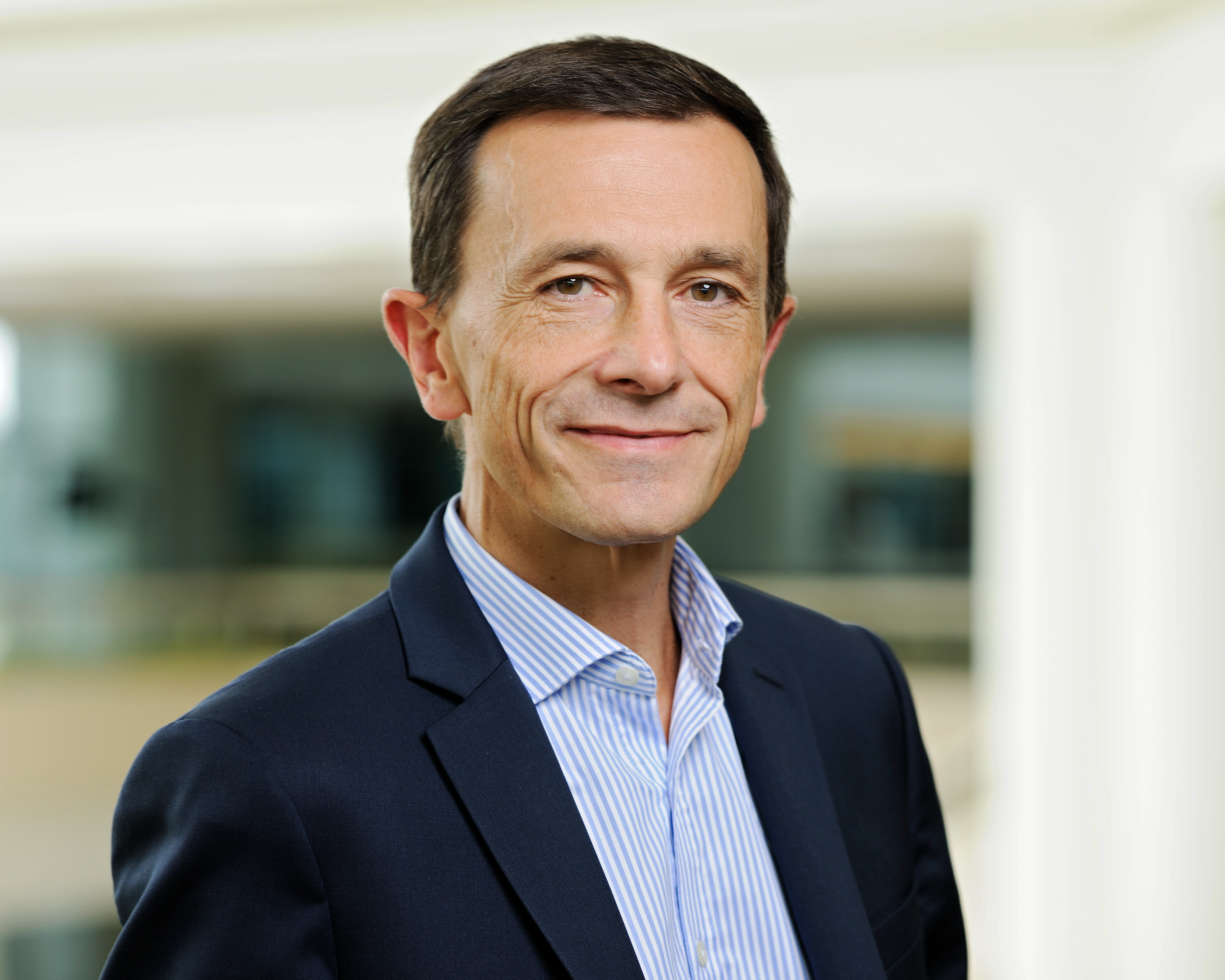 Portrait d'un homme d'âge mûr en blazer bleu et chemise à rayures, souriant dans un cadre professionnel.