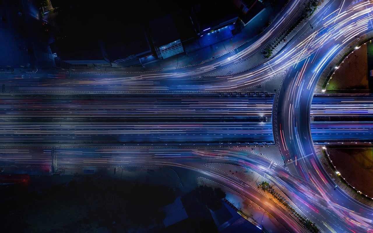 Vue aérienne de nuit d'un échangeur routier avec des traînées lumineuses de véhicules, symbolisant le mouvement et la connectivité urbaine.