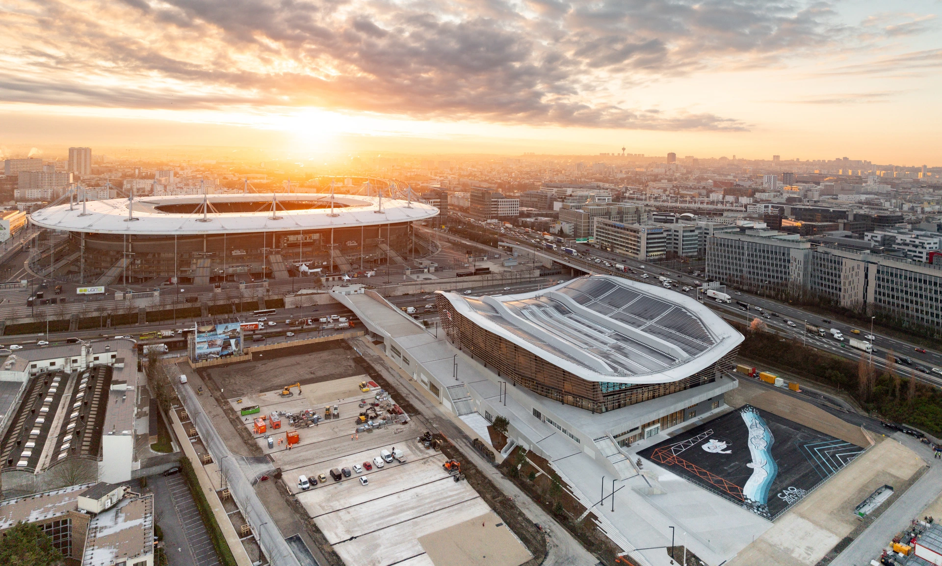 Vue aérienne d'un coucher de soleil flamboyant sur le Stade de France et le Centre Aquatique Olympique de Paris 2024, entourés d'autoroutes animées et d'un paysage urbain dense. Le soleil orange éclatant illumine les toits clairs des bâtiments et les nuages, contrastant avec les teintes grises de la ville.