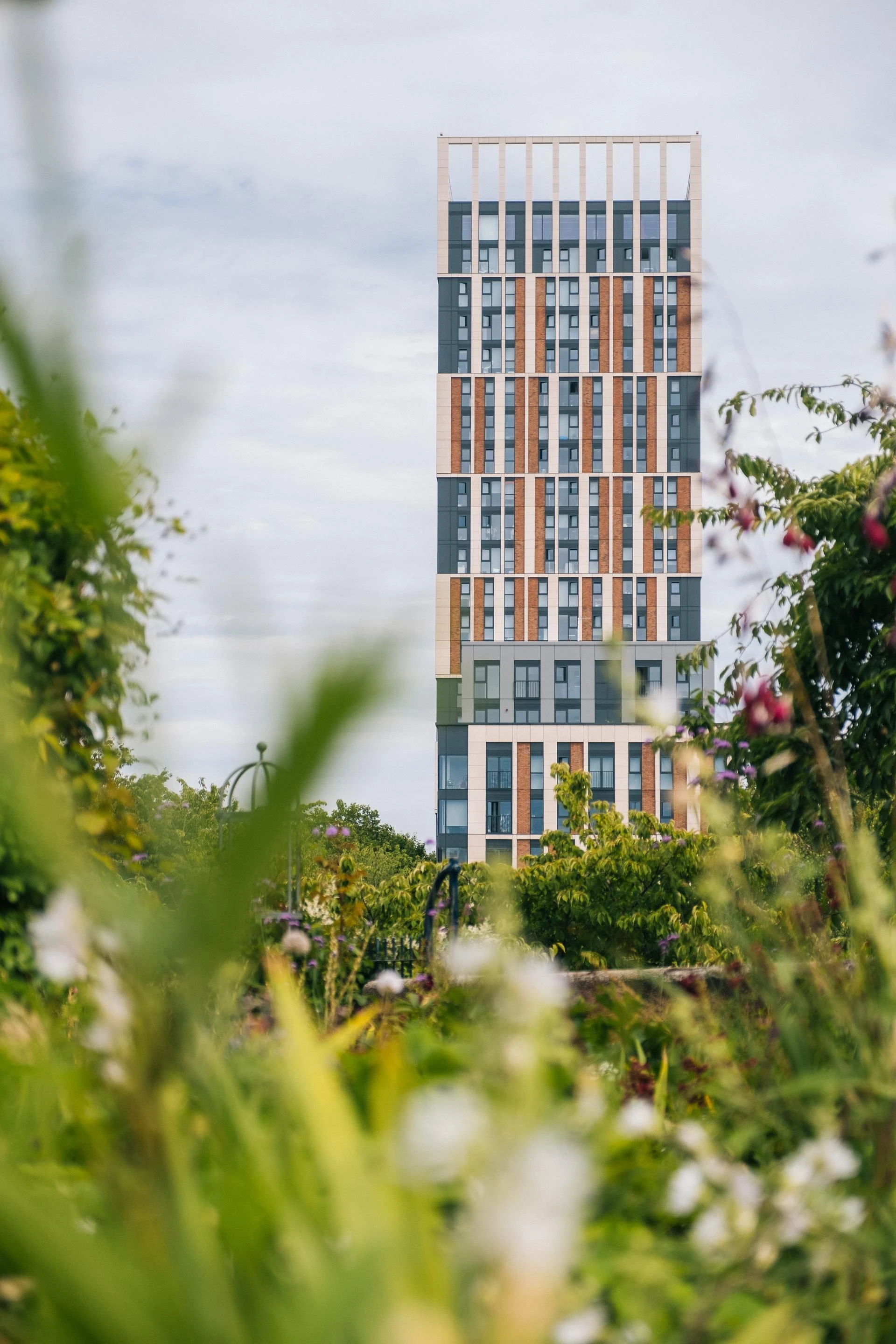 Un gratte-ciel moderne avec une façade à motifs verticaux gris foncé, crème et brun-rougeâtre, parsemée de nombreuses fenêtres, est visible au loin, encadré par une végétation verte floue et des fleurs au premier plan.