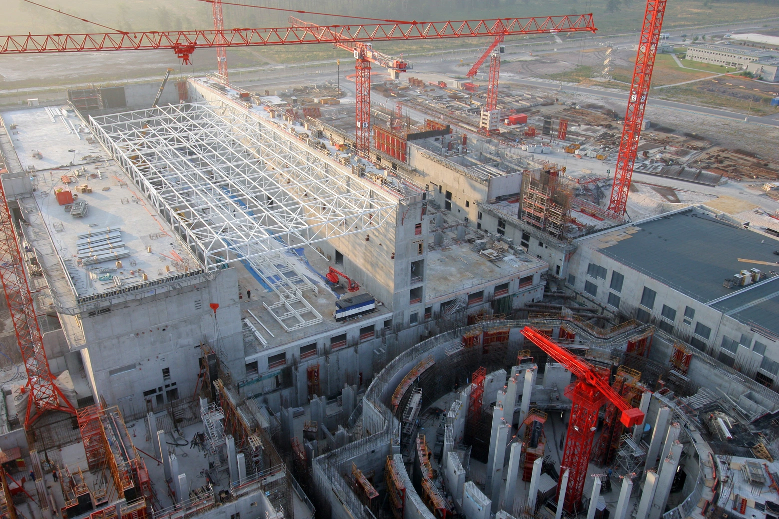 Vue aérienne d'un vaste chantier de construction en cours avec de multiples grues rouges, des bâtiments en béton gris et une grande structure de toit en treillis métallique blanc. Au premier plan se trouve une fosse de fondation circulaire profonde avec des piliers, suggérant un projet industriel majeur.