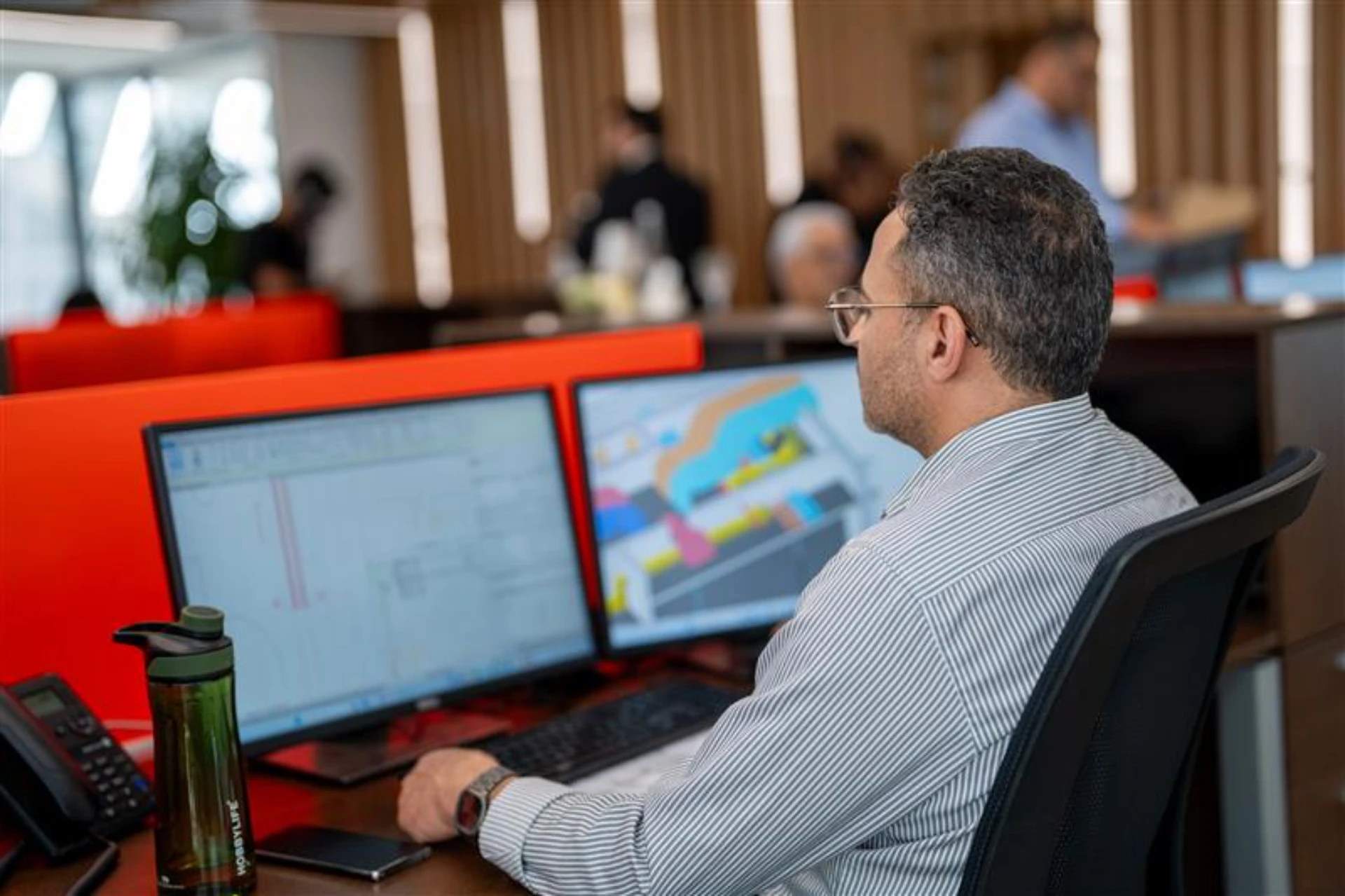 Un homme aux cheveux gris et lunettes, vêtu d'une chemise rayée, est assis de dos devant deux moniteurs d'ordinateur affichant des logiciels de conception architecturale ou d'ingénierie colorés dans un bureau moderne avec des séparations orange.