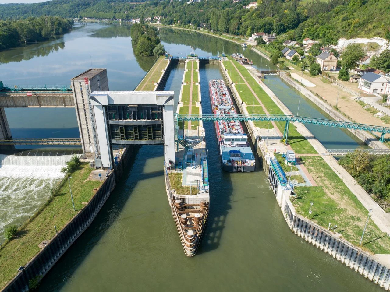 Vue aérienne d'un grand complexe d'écluses et d'un barrage avec une chute d'eau sur une rivière sinueuse. Deux péniches naviguent dans les bassins, entourés de rives verdoyantes, de structures grises et vertes, et de collines boisées sous un ciel clair.