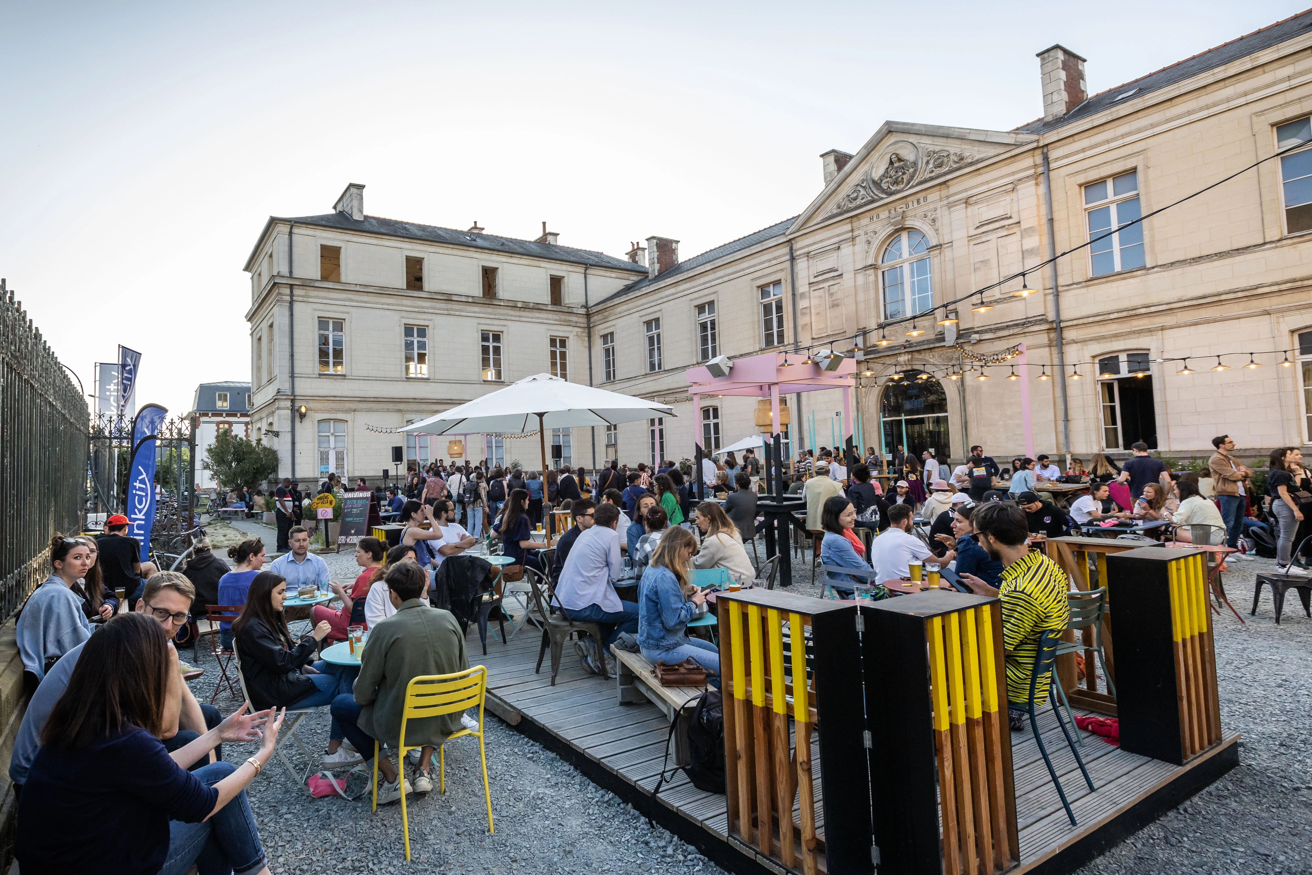 Une réunion animée en plein air rassemble de nombreuses personnes qui discutent autour de tables et de bancs installés sur des terrasses en bois et du gravier, sous des guirlandes lumineuses et un grand parasol blanc, le tout devant un imposant bâtiment historique de couleur claire.