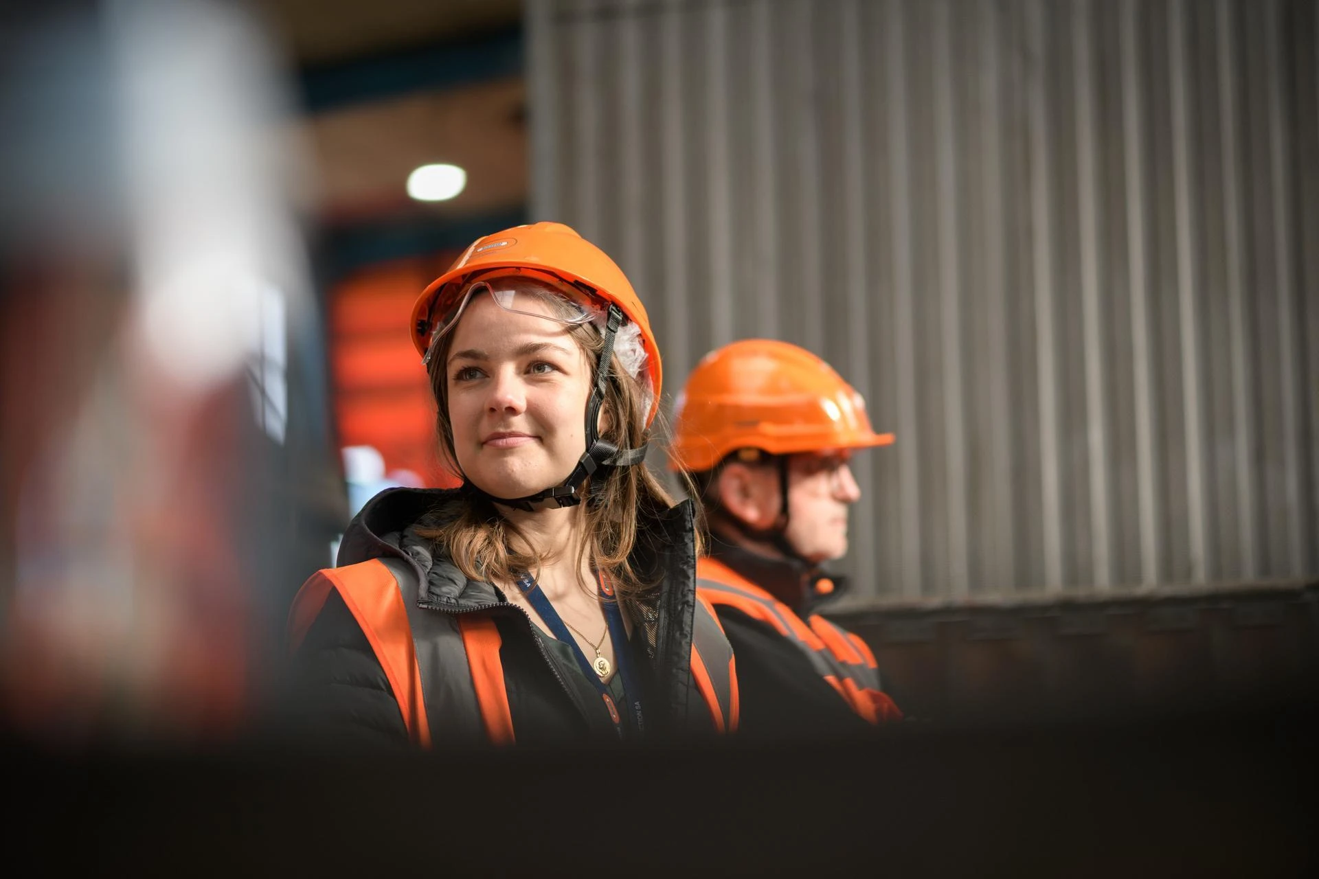 Une jeune femme souriante, portant un casque de sécurité orange, des lunettes de protection et un gilet réfléchissant, regarde vers la gauche dans un environnement industriel. Un autre travailleur en équipement similaire est flou en arrière-plan.