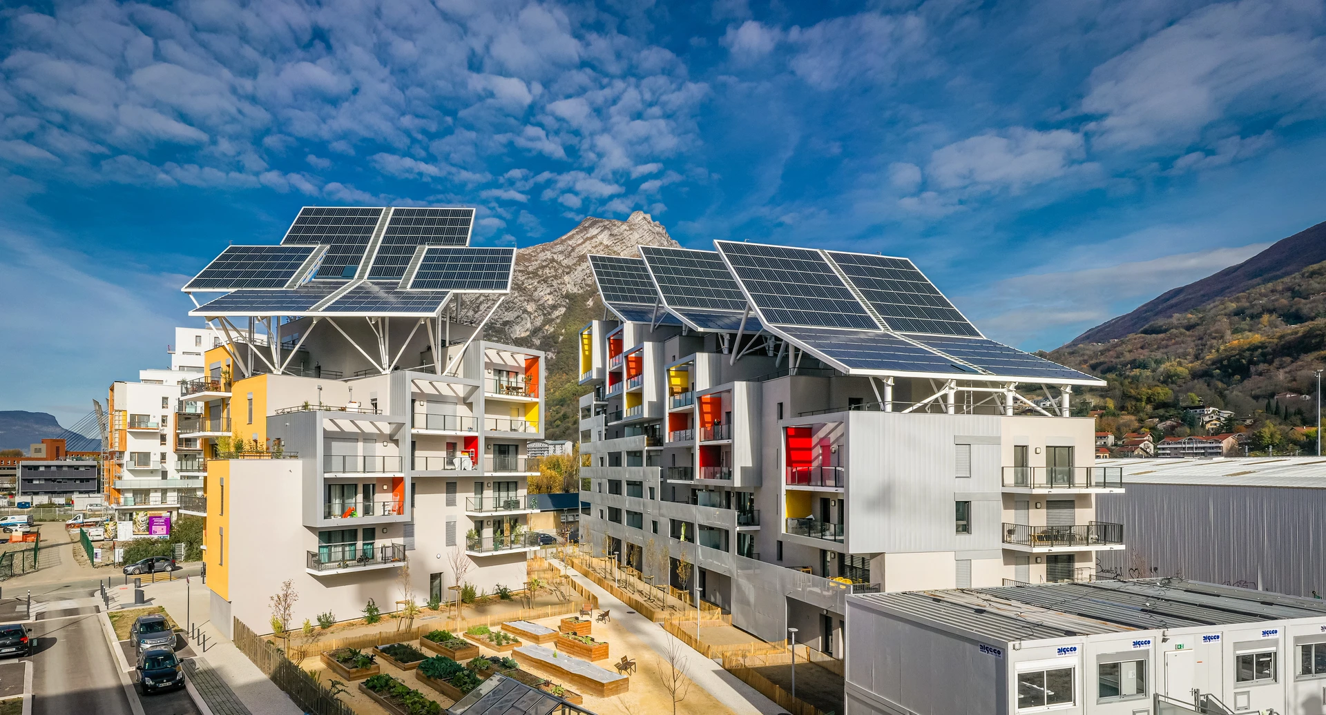 Vue panoramique de bâtiments résidentiels modernes avec de grands panneaux solaires inclinés sur leurs toits et des balcons colorés, sous un ciel bleu nuageux avec des montagnes boisées en arrière-plan.
