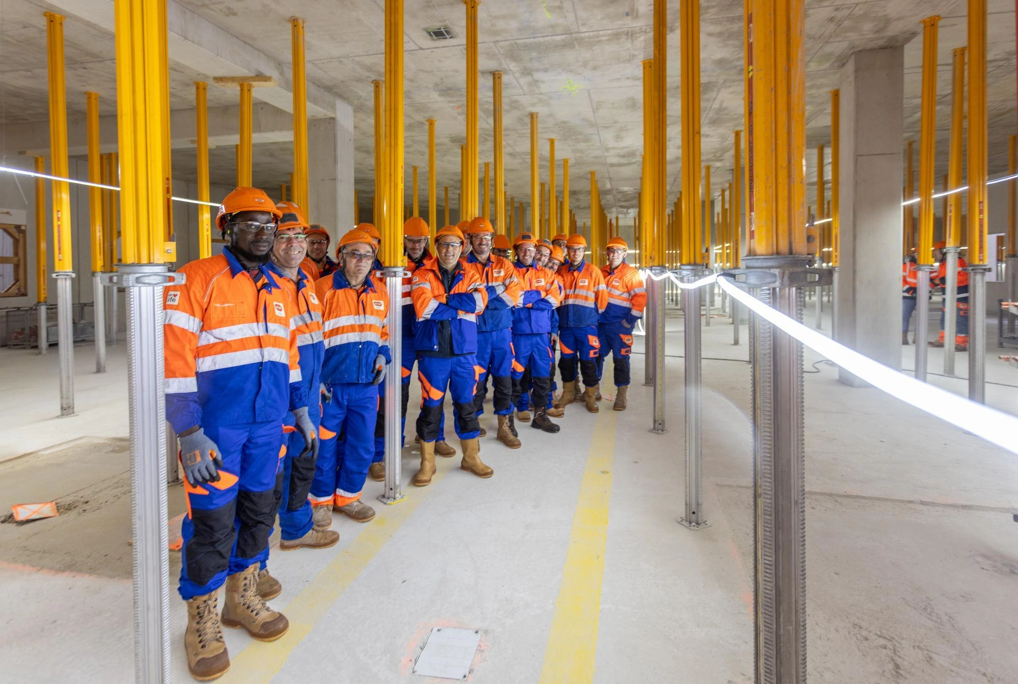 Un groupe d'ouvriers du bâtiment souriants, vêtus d'uniformes de travail orange et bleu vif et de casques orange, posent dans une structure en béton en construction, caractérisée par de nombreux piliers de soutien jaunes.