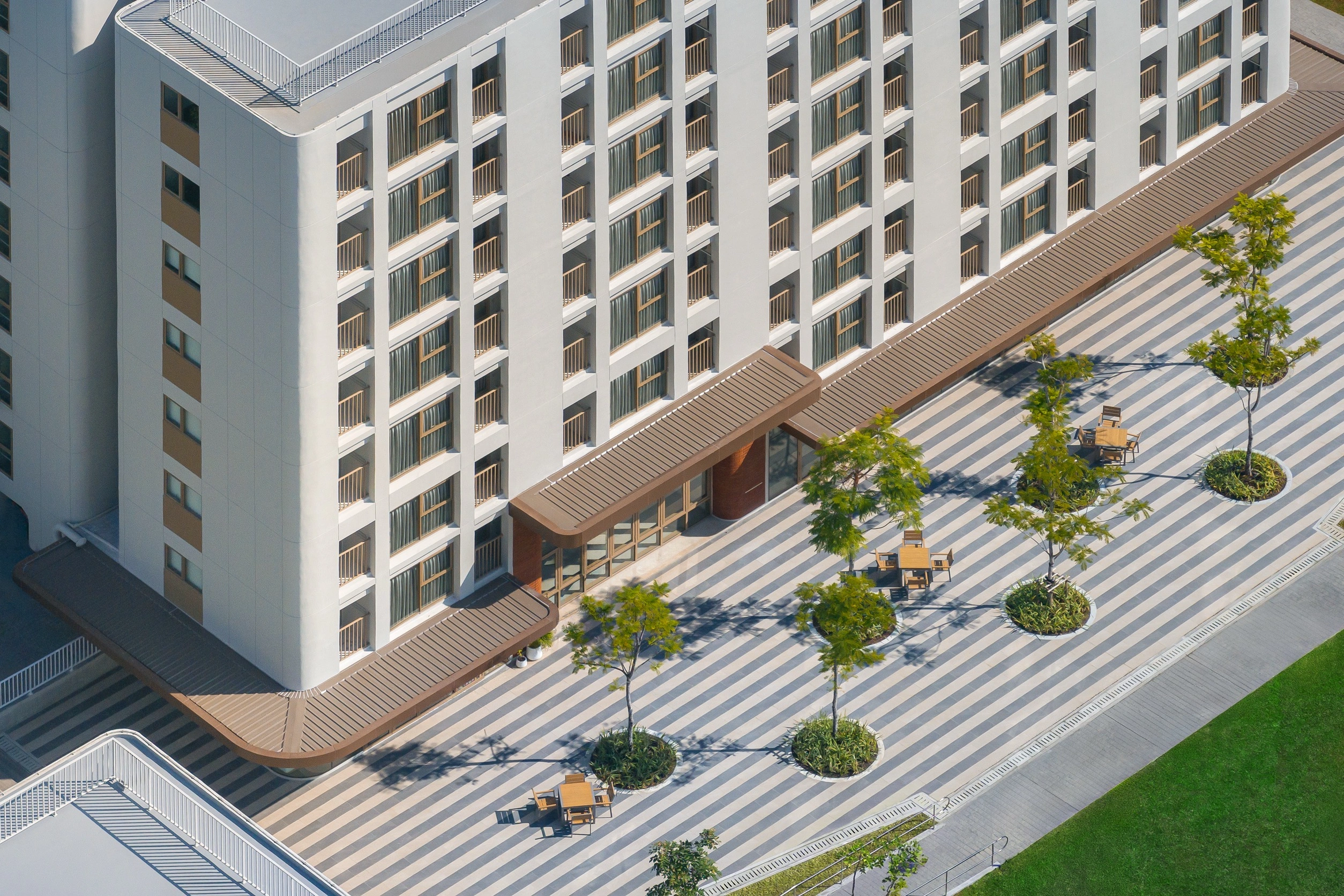 Vue aérienne d'un bâtiment blanc moderne et d'un patio rayé avec arbres et mobilier extérieur.