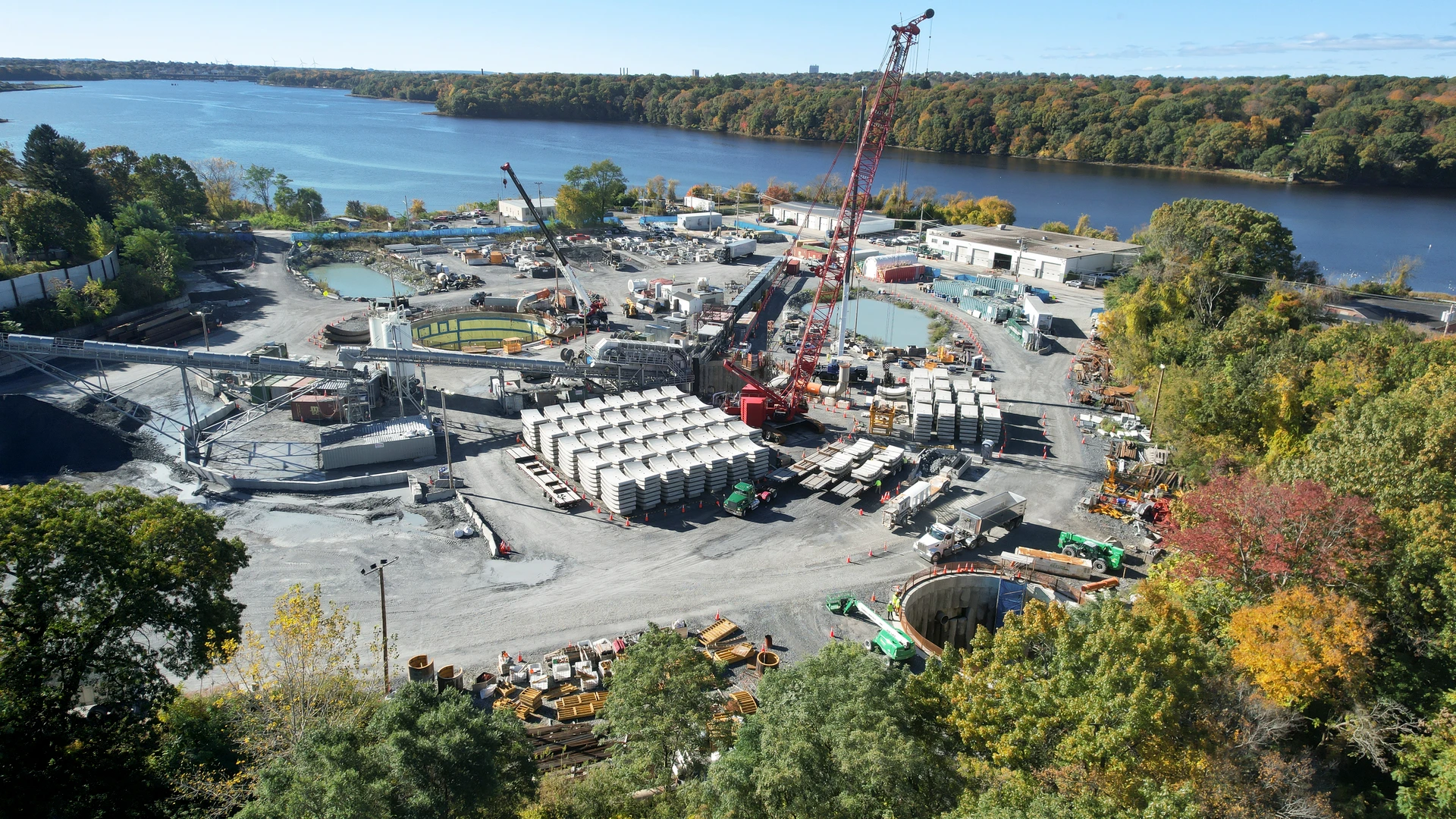 An aerial view reveals a vast industrial construction site next to a blue river, featuring a large red crane, conveyor systems, concrete processing tanks, and numerous stacks of grey precast segments, all framed by vibrant green, yellow, and red autumn trees under a clear sky.