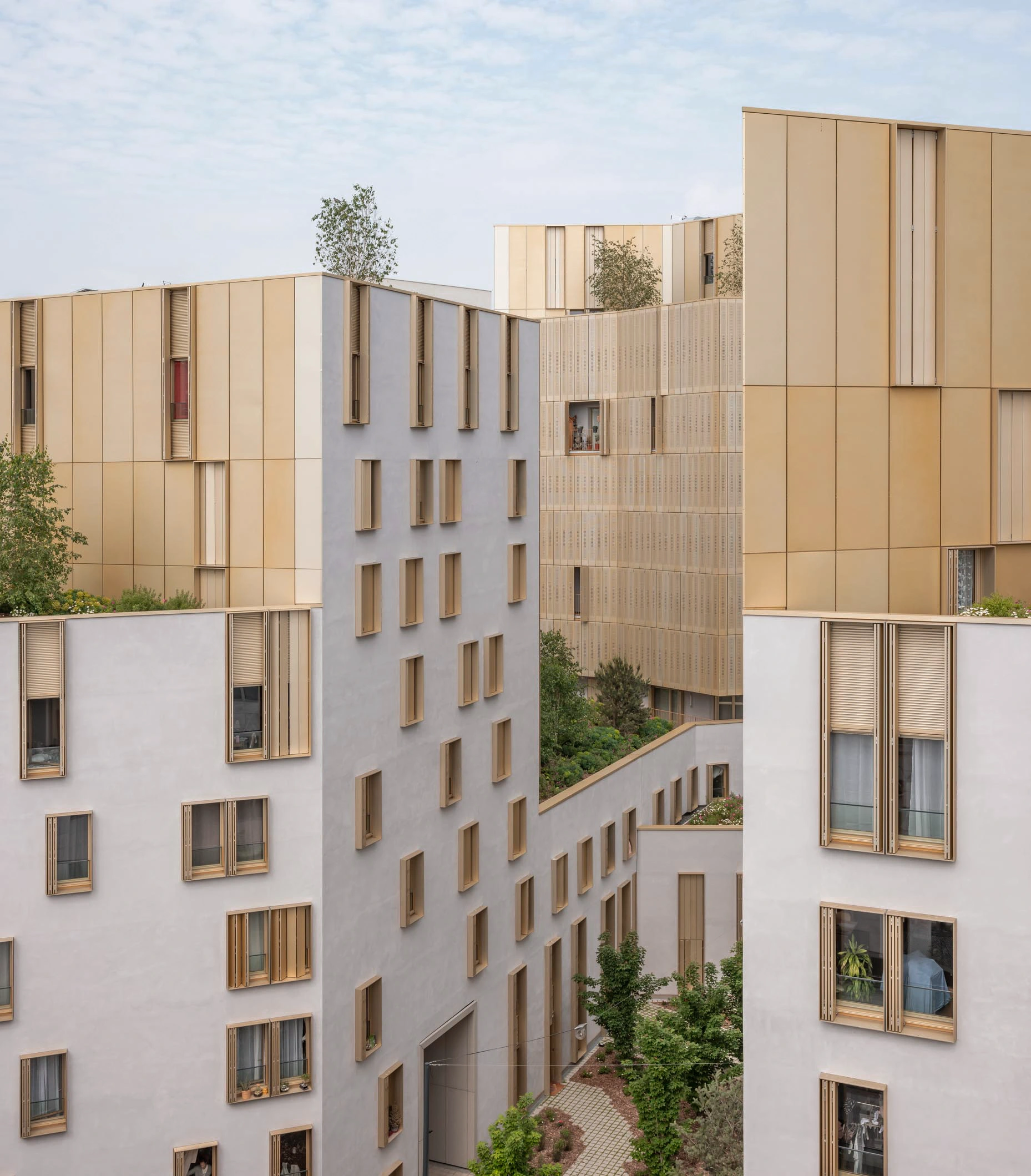 Un complexe résidentiel moderne présente des bâtiments aux façades combinant des panneaux dorés brillants et des murs gris clair, intégrant des toits végétalisés et des espaces verts en cour intérieure sous un ciel nuageux.