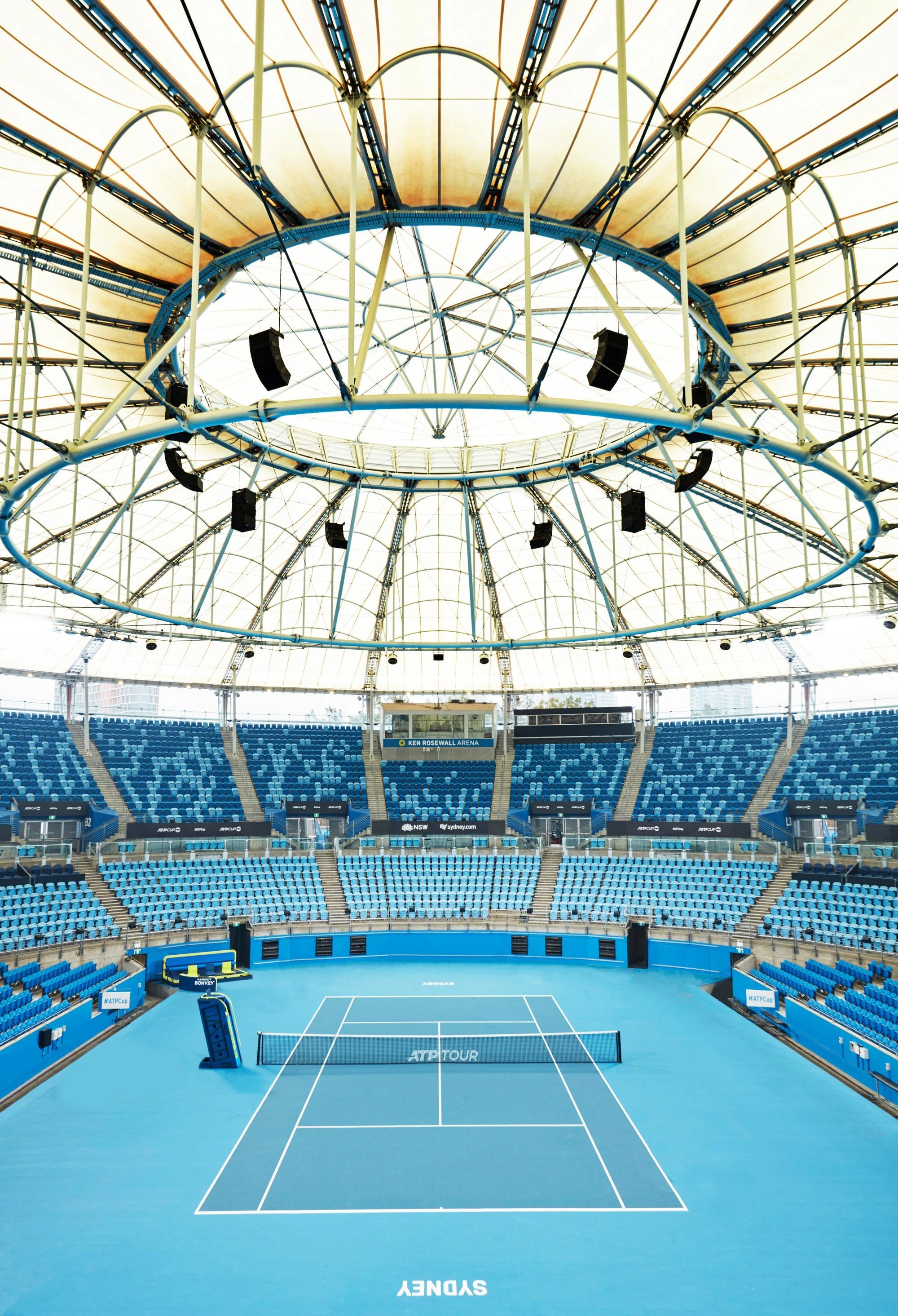 Vue aérienne d'un court de tennis bleu vif et vide dans le stade Ken Rosewall Arena, avec un filet ATP Tour et des rangées de sièges bleus clairs. Le stade est recouvert d'une grande structure de toit en toile blanche et bleue.
