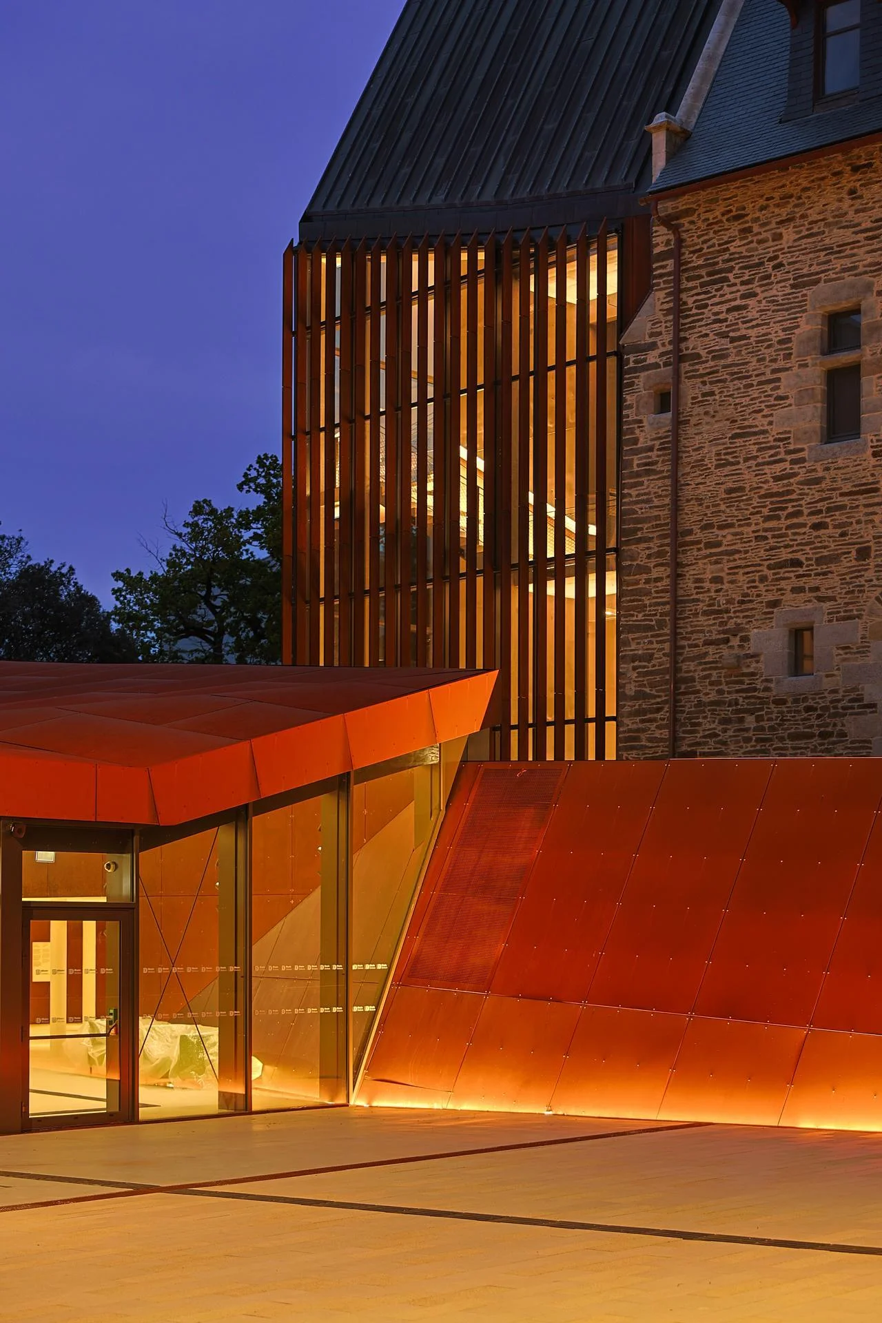 Façade d'un bâtiment moderne en panneaux orange-rouge et lattes verticales illuminées, adossé à une structure en pierre ancienne, sous un ciel crépusculaire.