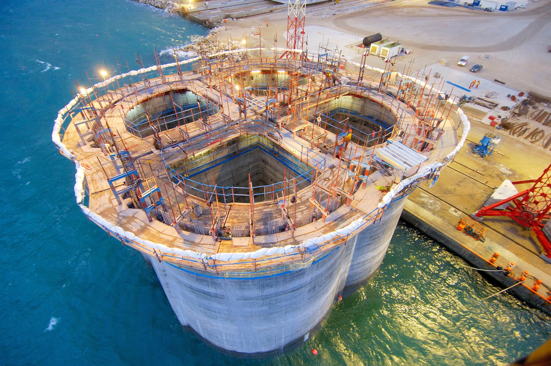 Vue aérienne d'une vaste structure en béton multi-lobée en construction dans une eau bleu-vert, avec des échafaudages, des puits circulaires profonds et un éclairage intense, à côté d'un quai.