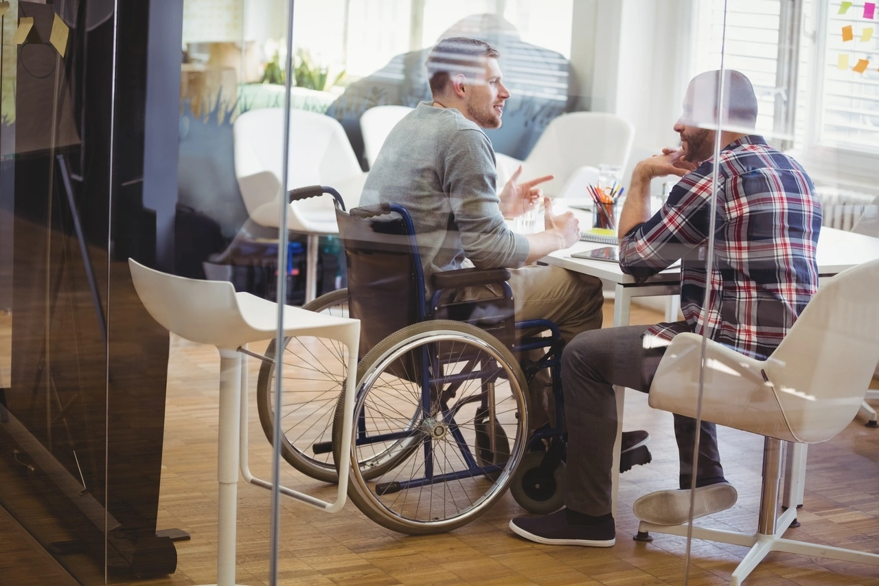 Deux hommes sont engagés dans une discussion à une table blanche dans un bureau moderne ; l'un, en fauteuil roulant bleu, gesticule tandis que l'autre l'écoute attentivement. La scène, vue à travers une cloison vitrée, illustre une interaction professionnelle et inclusive.