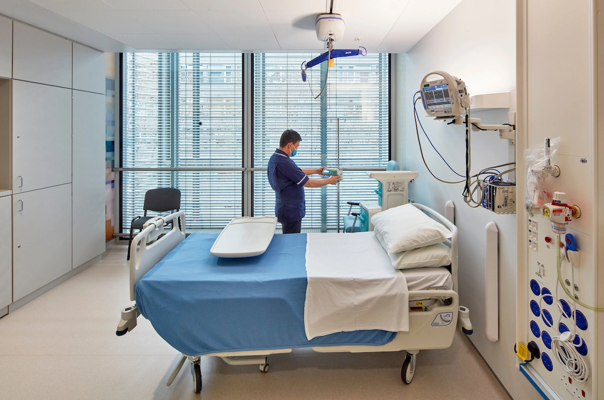 Une infirmière en tenue bleue et masque ajuste un dispositif médical près d'un lit d'hôpital aux draps bleus et blancs, dans une chambre moderne et lumineuse avec de grandes fenêtres.