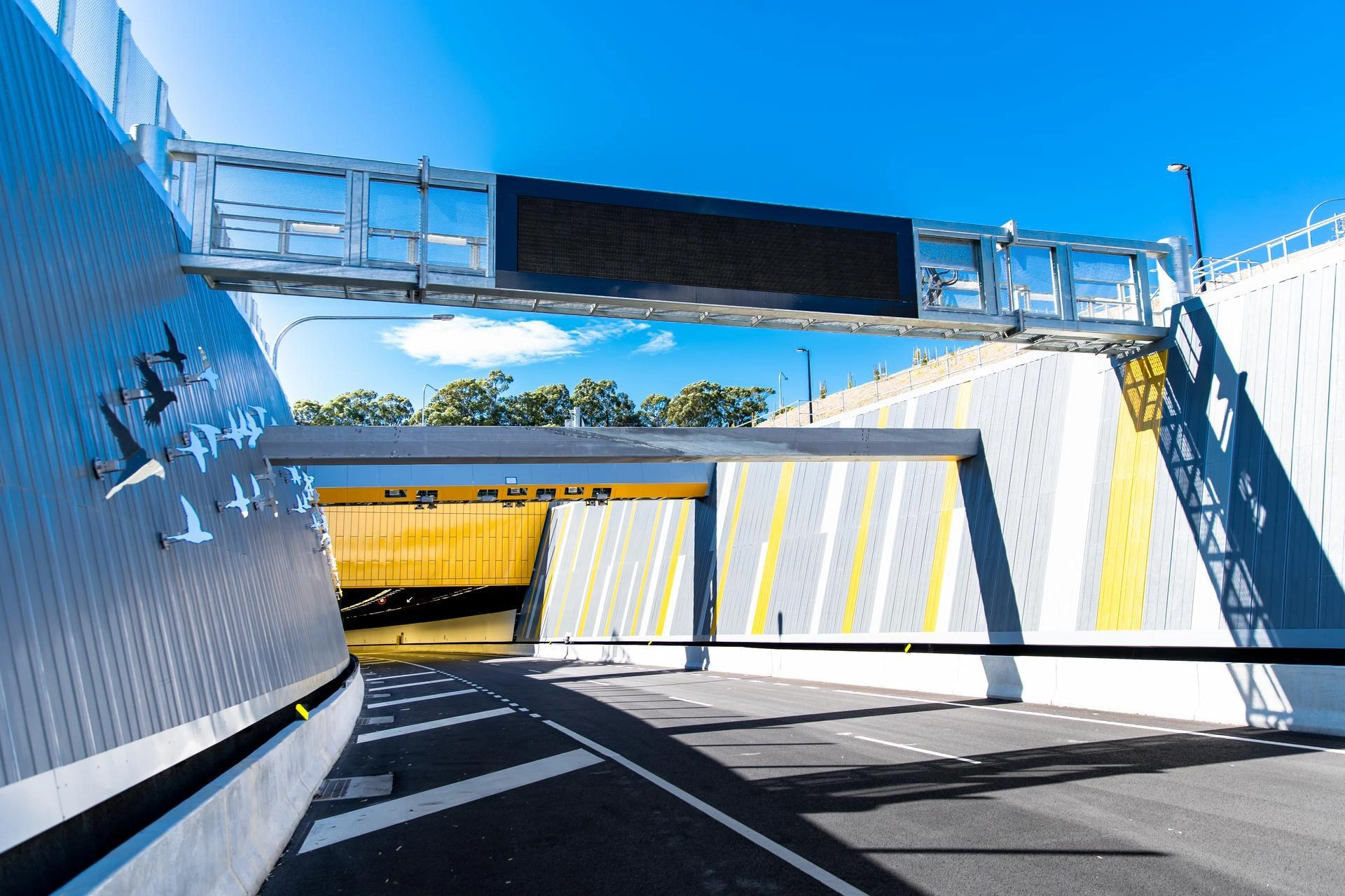 Entrée d'un tunnel autoroutier moderne sous un ciel bleu clair, avec des murs gris ornés de silhouettes d'oiseaux sur la gauche et de rayures verticales jaunes et blanches sur la droite, surmontés d'un grand panneau d'affichage électronique.