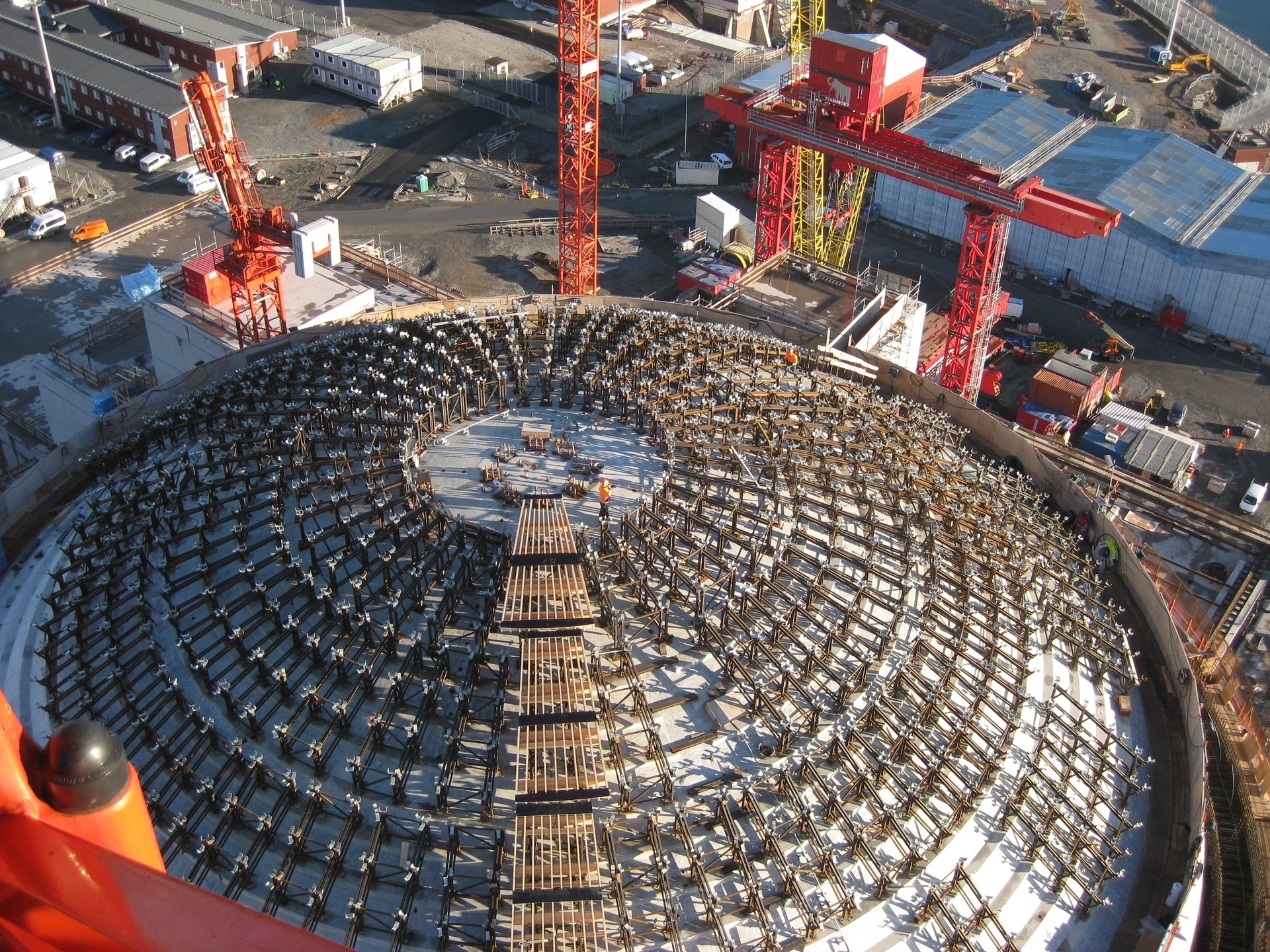 Vue aérienne d'une immense structure en dôme en construction, présentant un treillis métallique complexe disposé en cercles concentriques sur une base en béton gris, encadrée par de grandes grues rouges et jaunes sur un vaste chantier industriel ensoleillé.