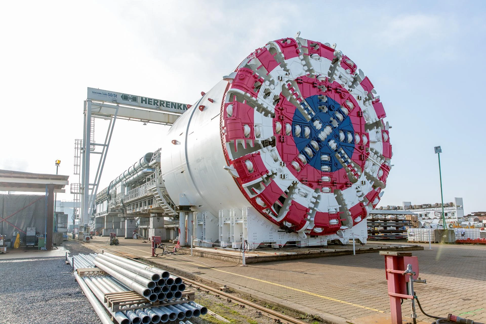 Une énorme machine de forage de tunnels (TBM) blanche, avec une tête de coupe rouge, blanche et bleue au premier plan, est préparée sur un site industriel. Un grand portique "HERRENKNECHT" est visible en arrière-plan sous un ciel dégagé.